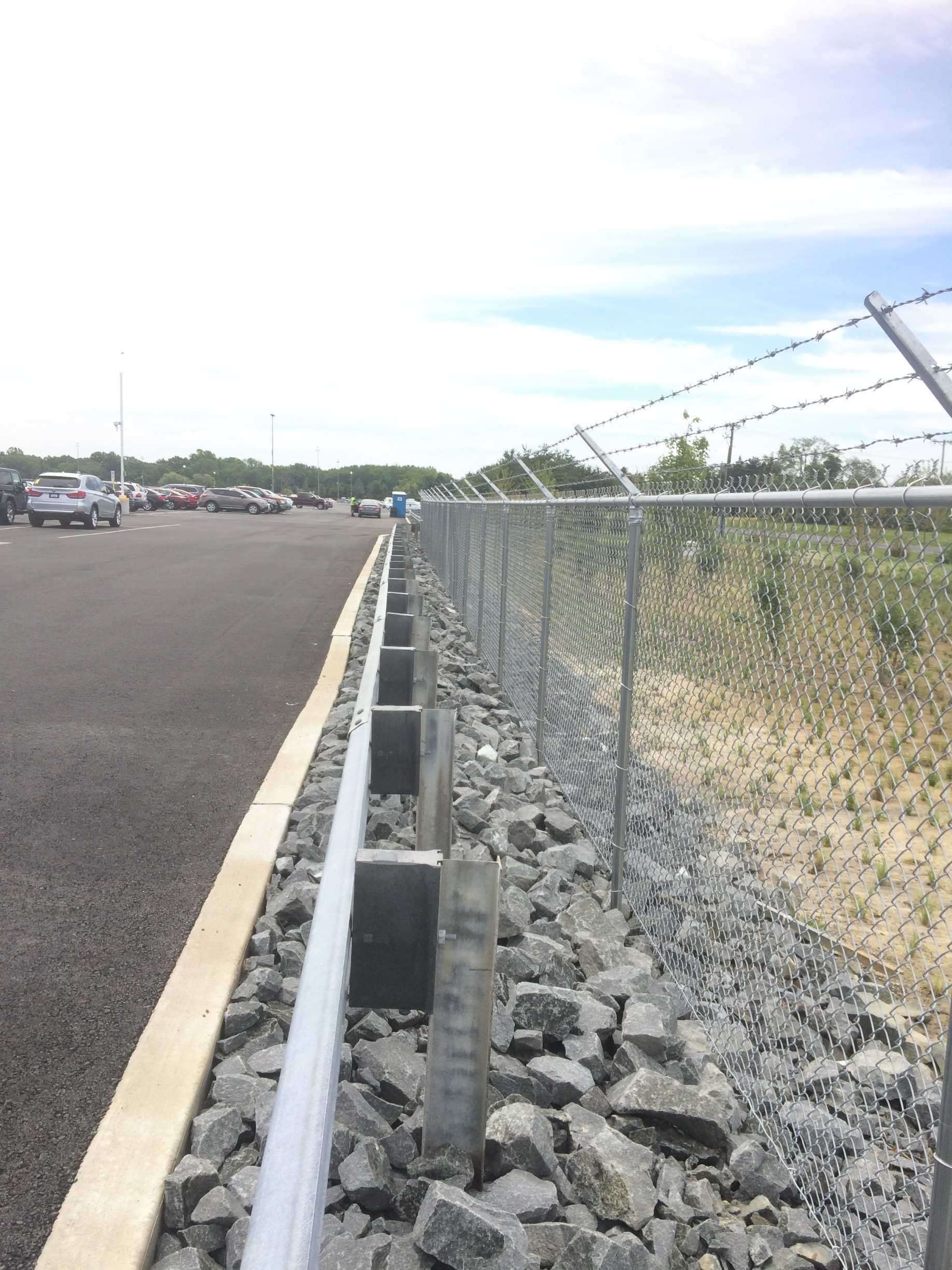 Chain-link fence with barbed wire atop, separating a parking lot from a gravel area and concrete drainage.