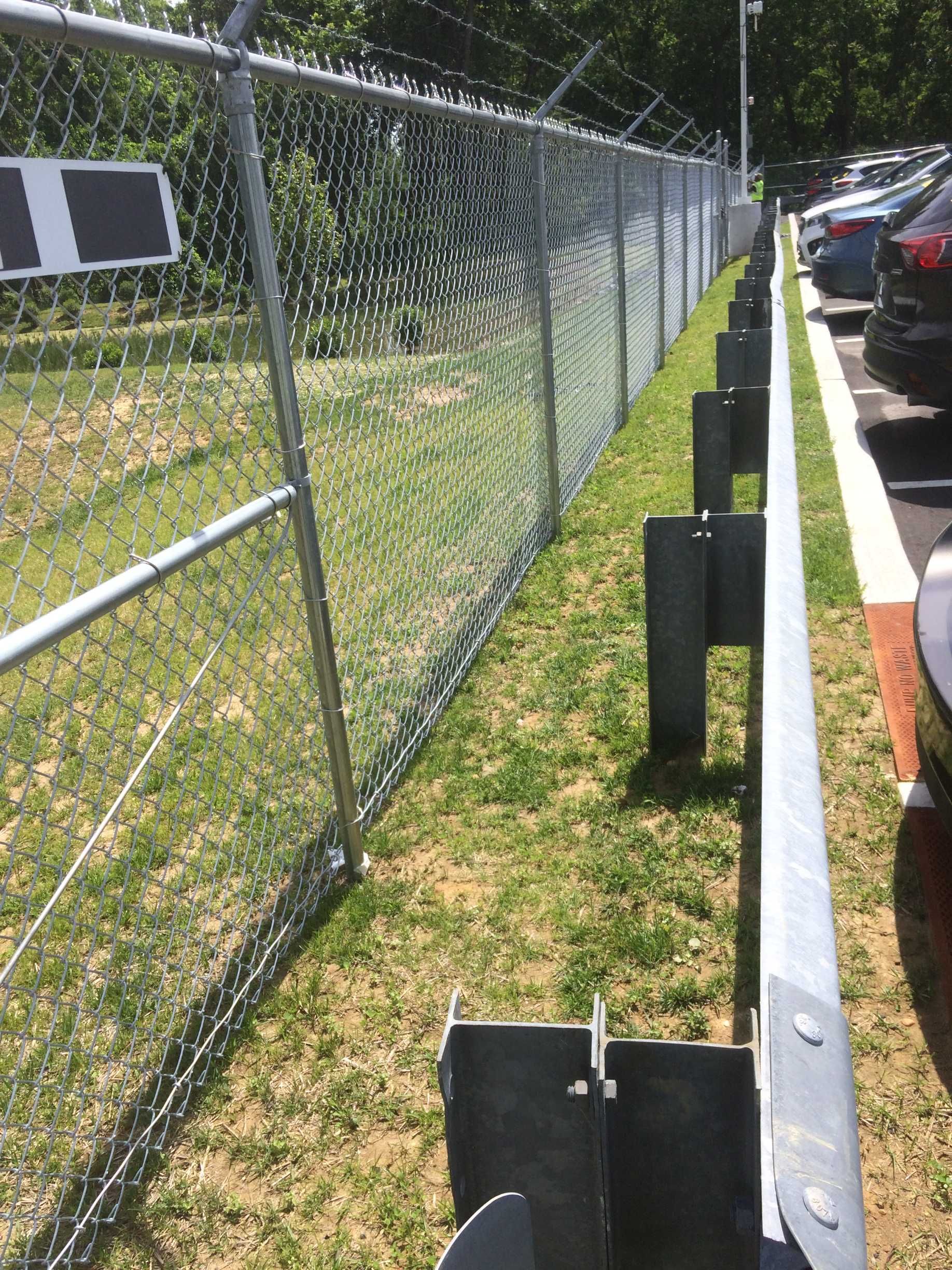 Chain-link fence with barbed wire atop, bordering a grassy area and parking lot with cars.