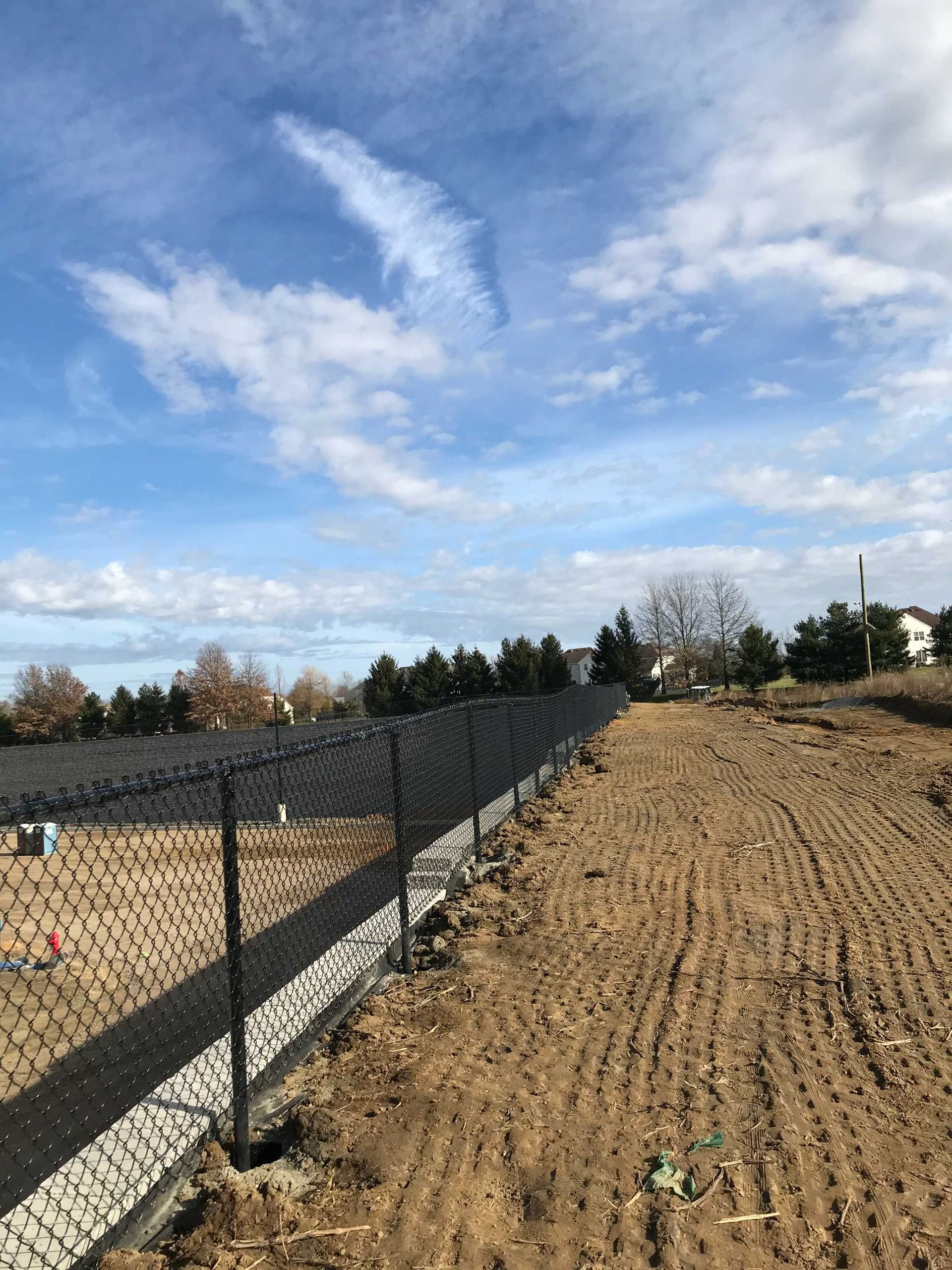 Chain link fence along a tilled dirt field under a cloudy blue sky.