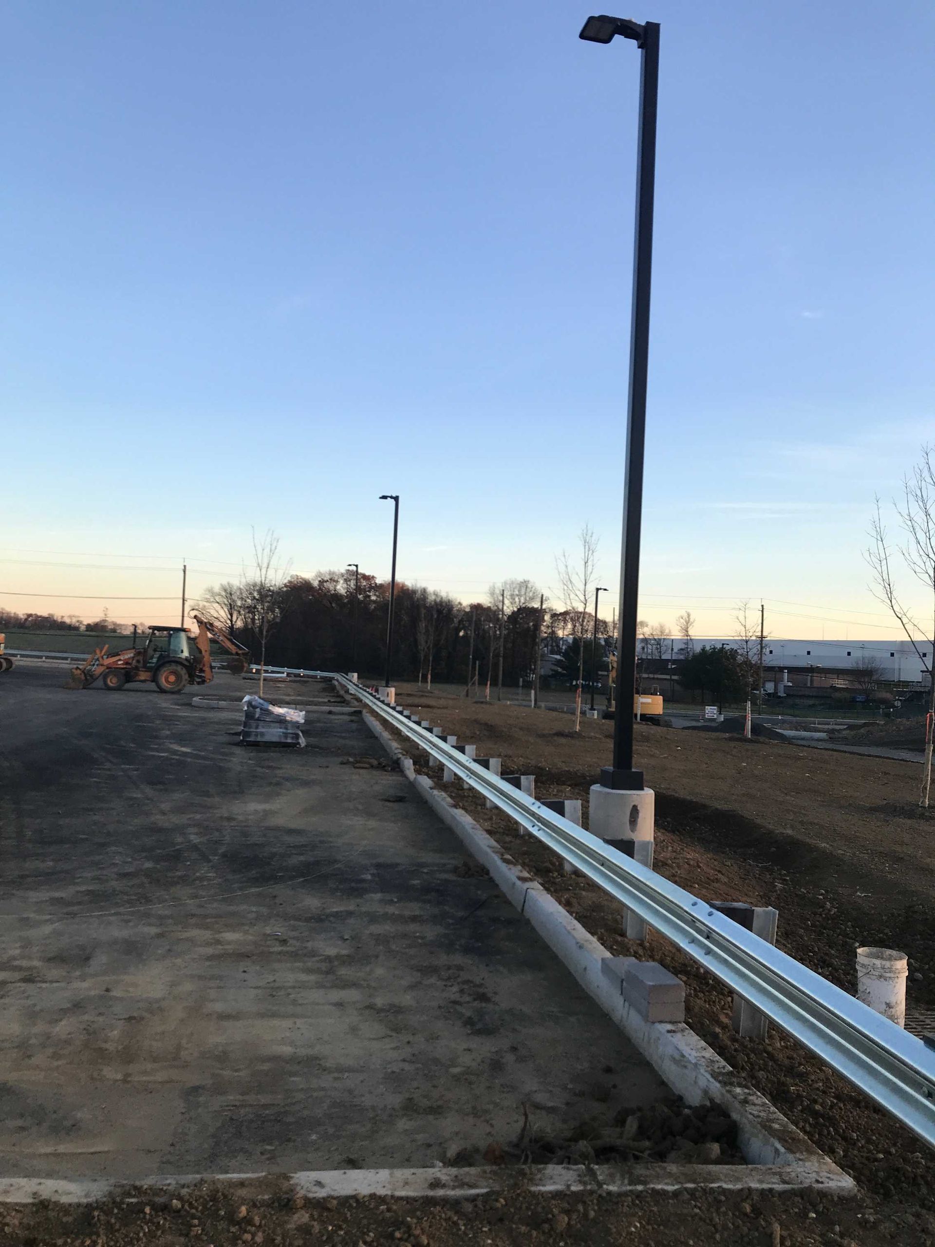 Parking lot construction: metal guardrail, tall light poles, bare trees, and machinery under a blue sky.