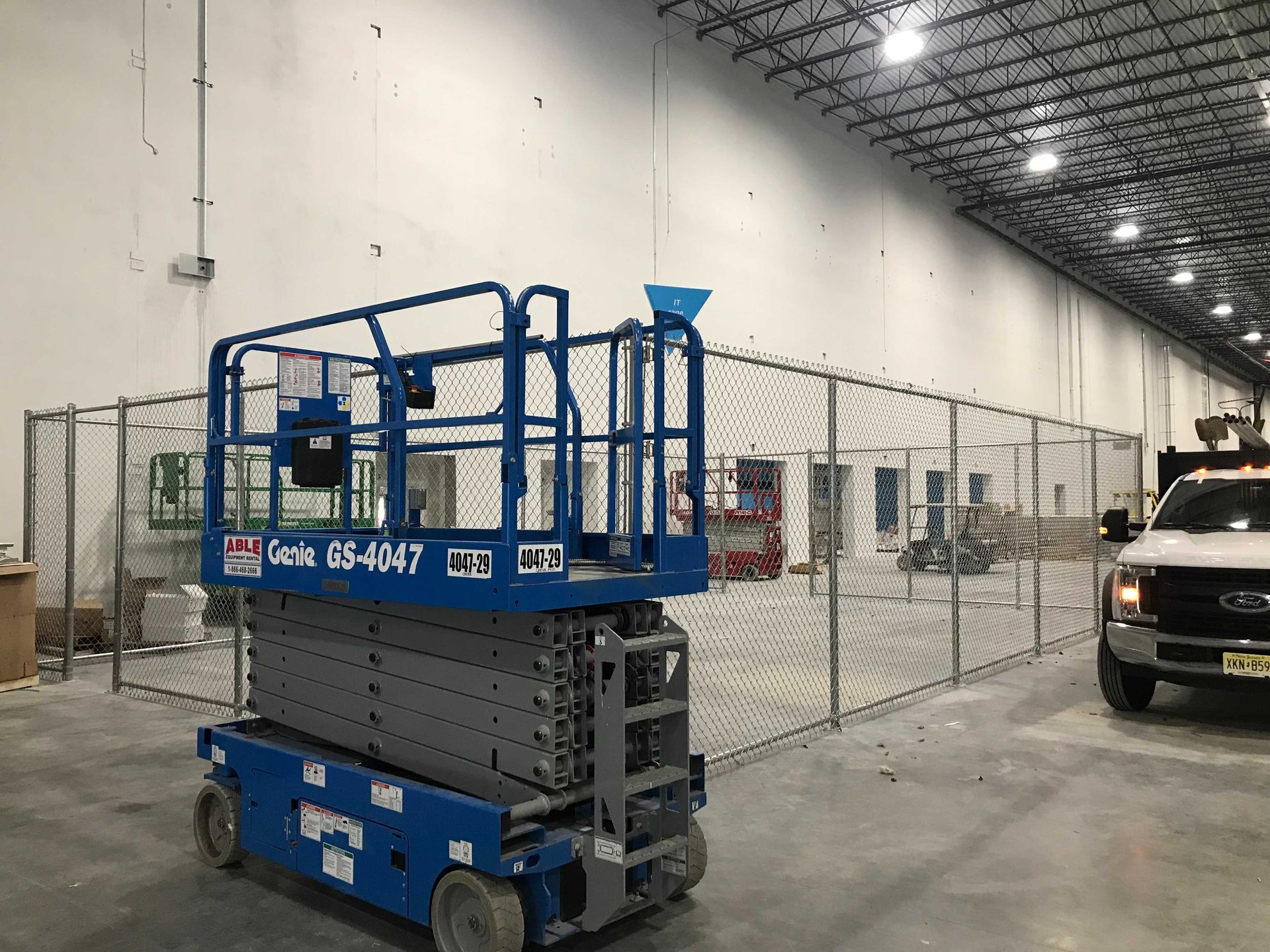 Blue scissor lift in an industrial space; a chain-link fence surrounds a construction area.