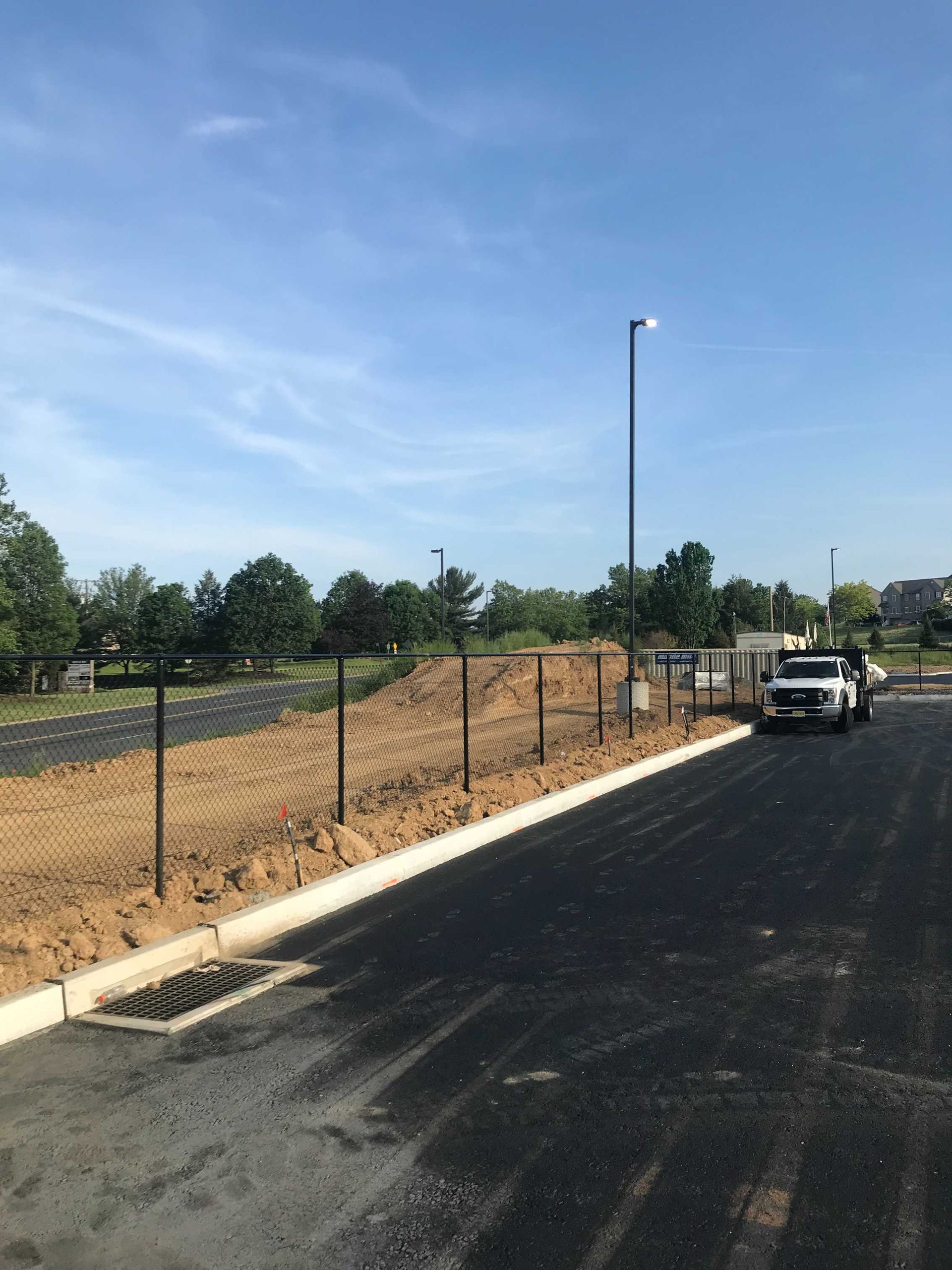 Black asphalt road and curb, with a chain-link fence, a pickup truck, and a blue sky in the background.