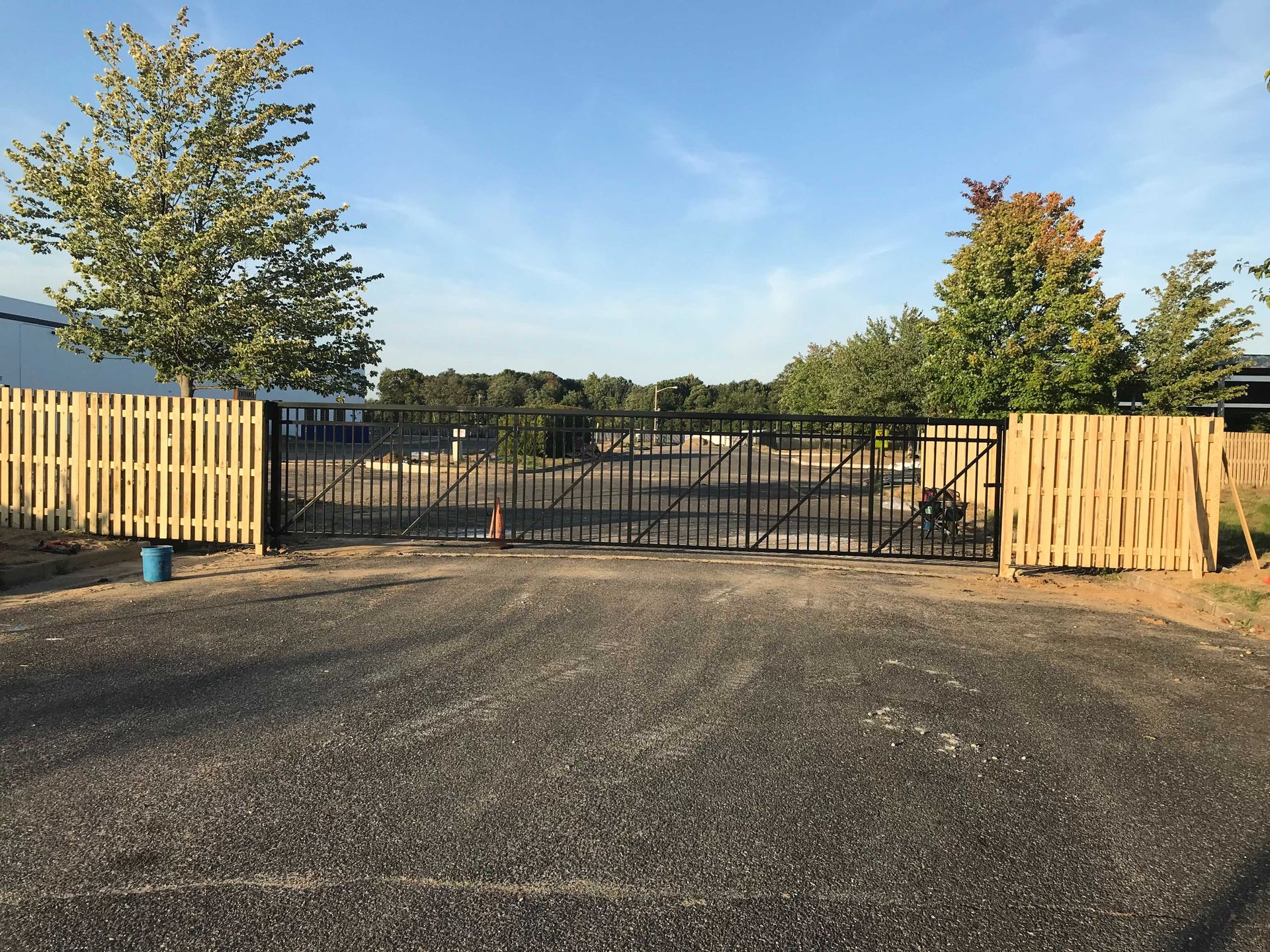 Black metal gate, flanked by wooden fences, opens onto a gravel driveway. Trees and sky are in the background.