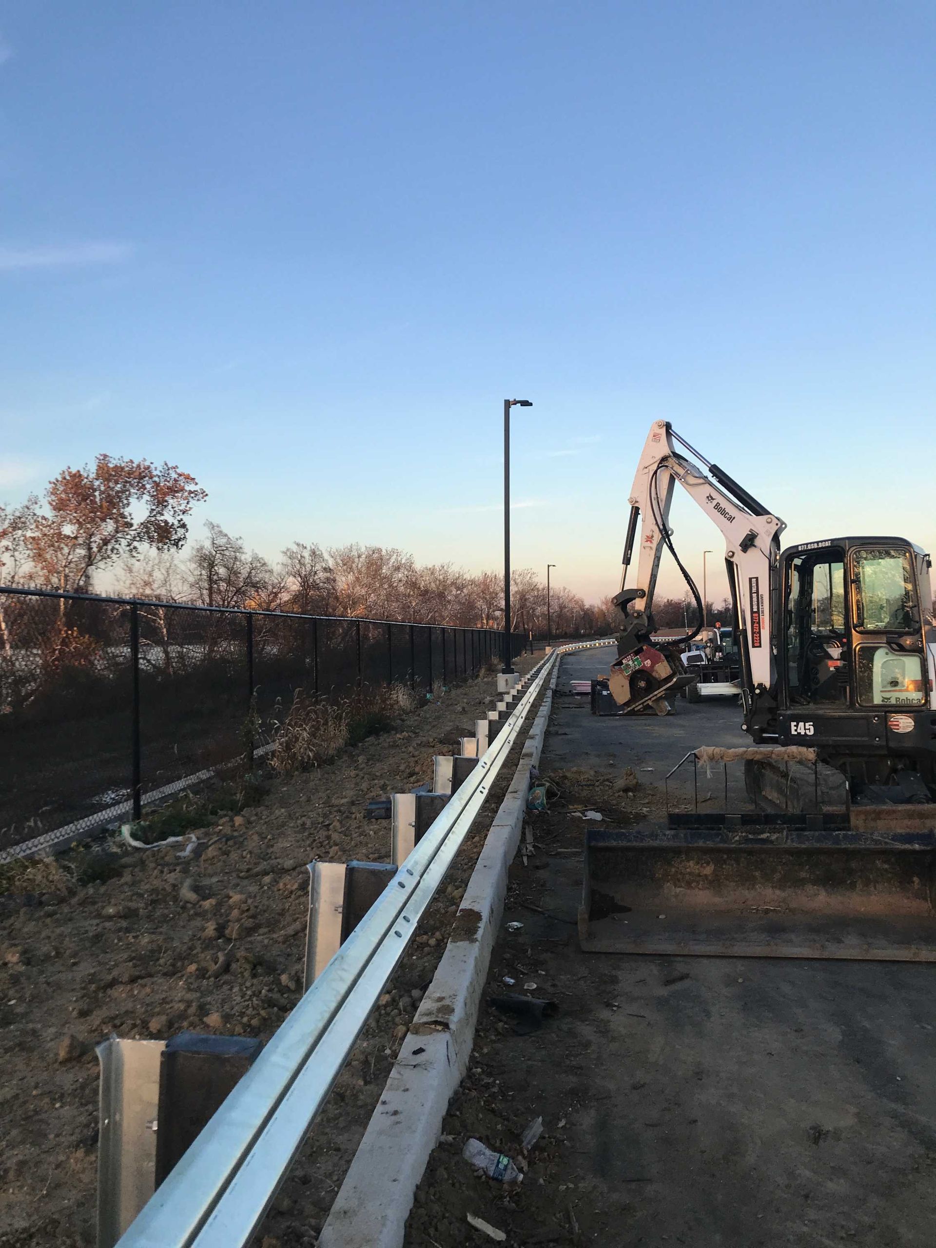 An excavator works next to a roadside barrier and dark metal poles under a blue sky.