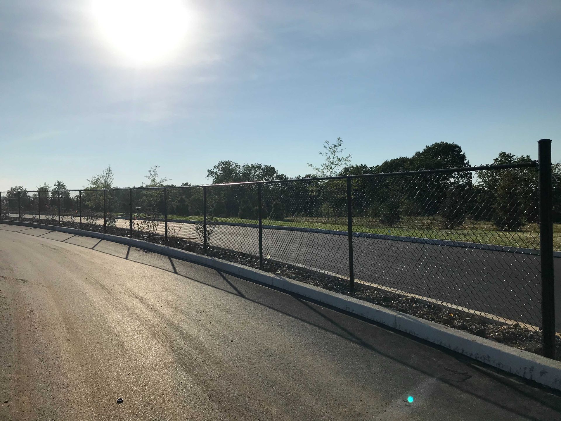 Black chain-link fence along a road, trees in the background, sunny sky overhead.