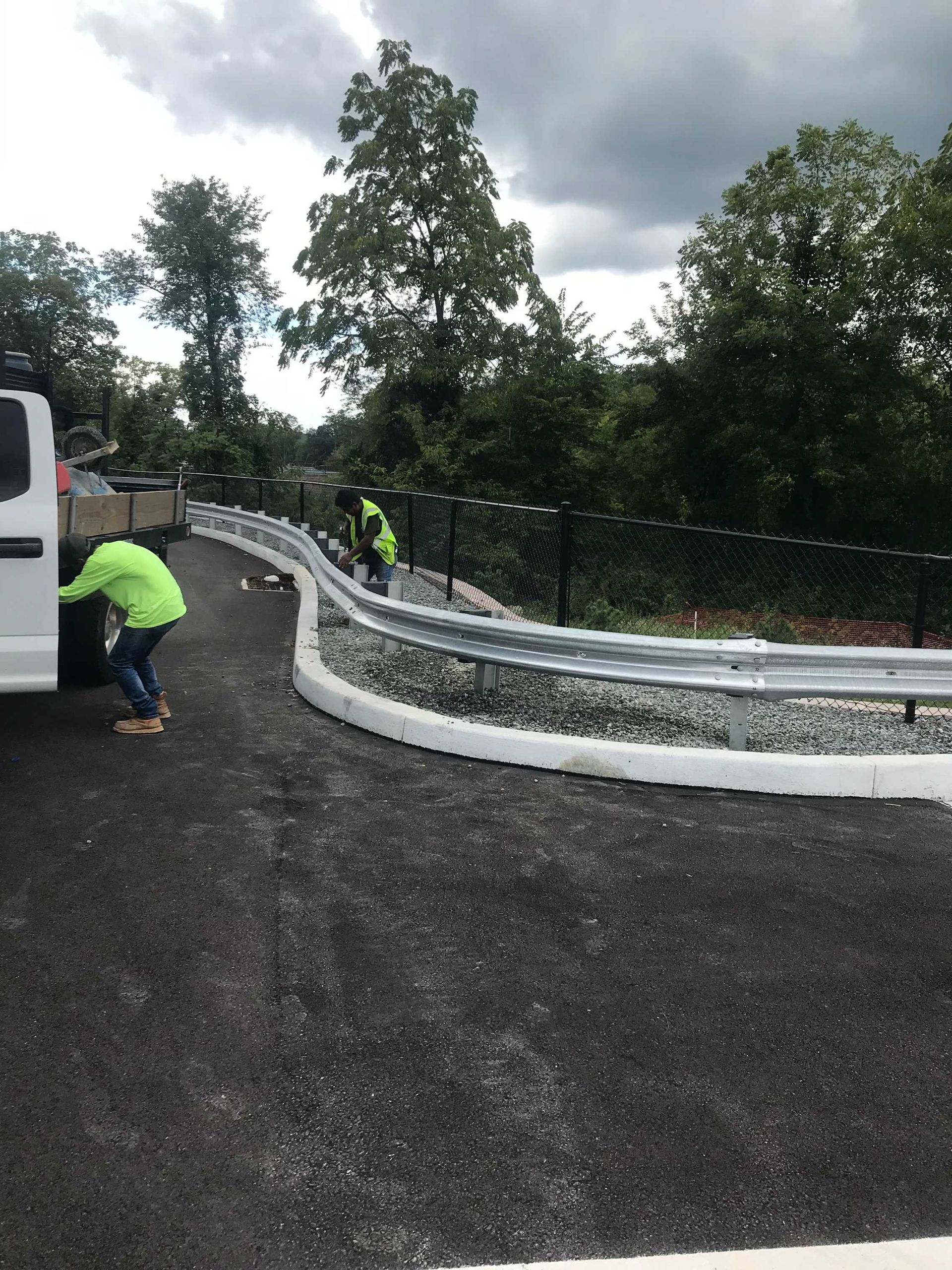 Workers install guardrail along a newly paved road. White van parked, green safety vests, gray sky.