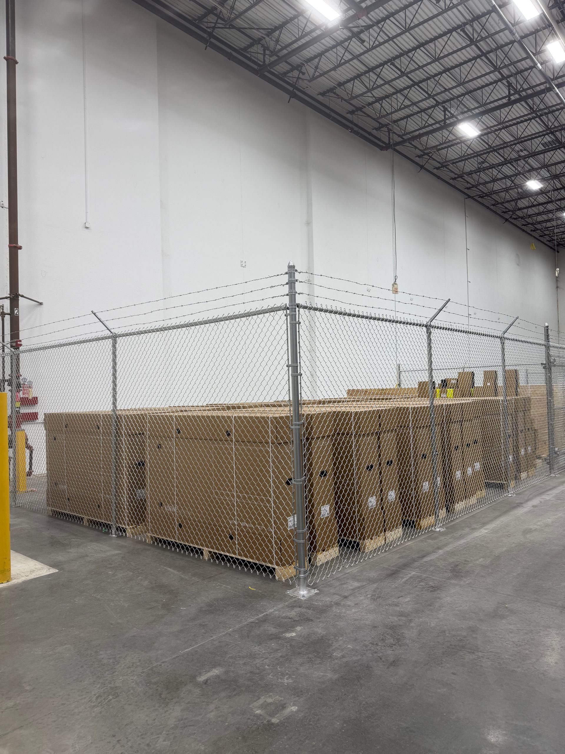 Stacked cardboard boxes are stored in an indoor warehouse behind a chain-link security fence with barbed wire.