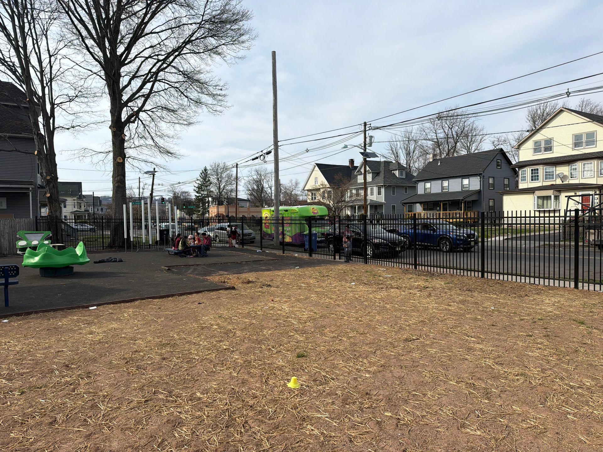 A neighborhood playground with wood chips, a green circular spinner, and a black metal fence with houses in the background.