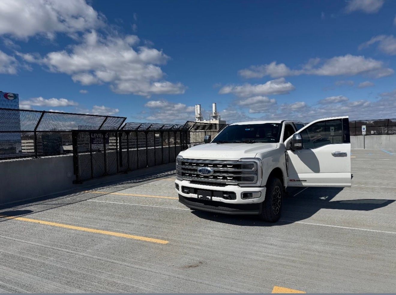 A white Ford pickup truck with its driver-side door open, parked on an outdoor rooftop parking deck under a blue sky.