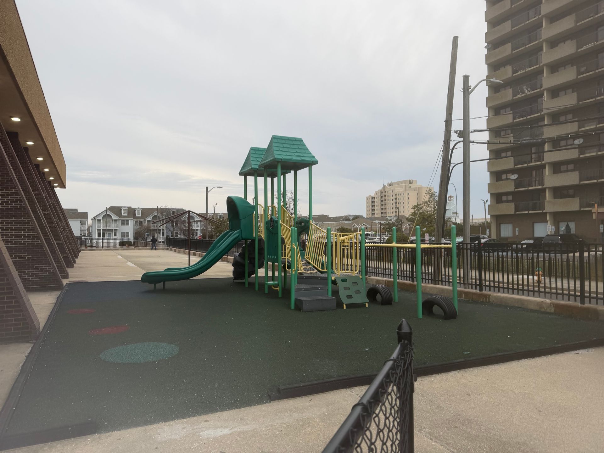 A green and yellow playground structure with a slide sits on a black rubberized surface next to a building and fence.