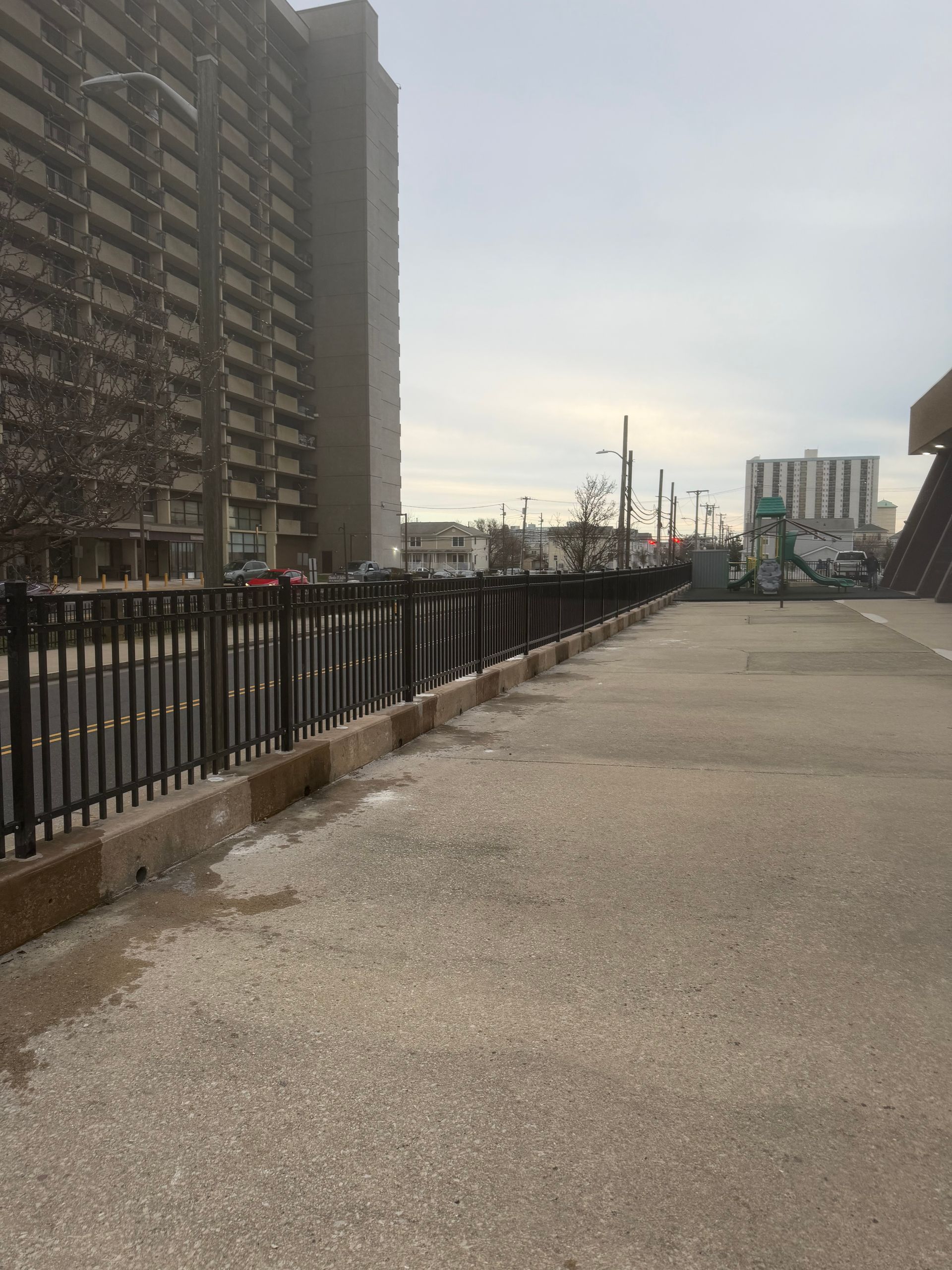 A wide paved walkway beside a black metal fence, with a tall apartment building on the left and city skyline in distance.