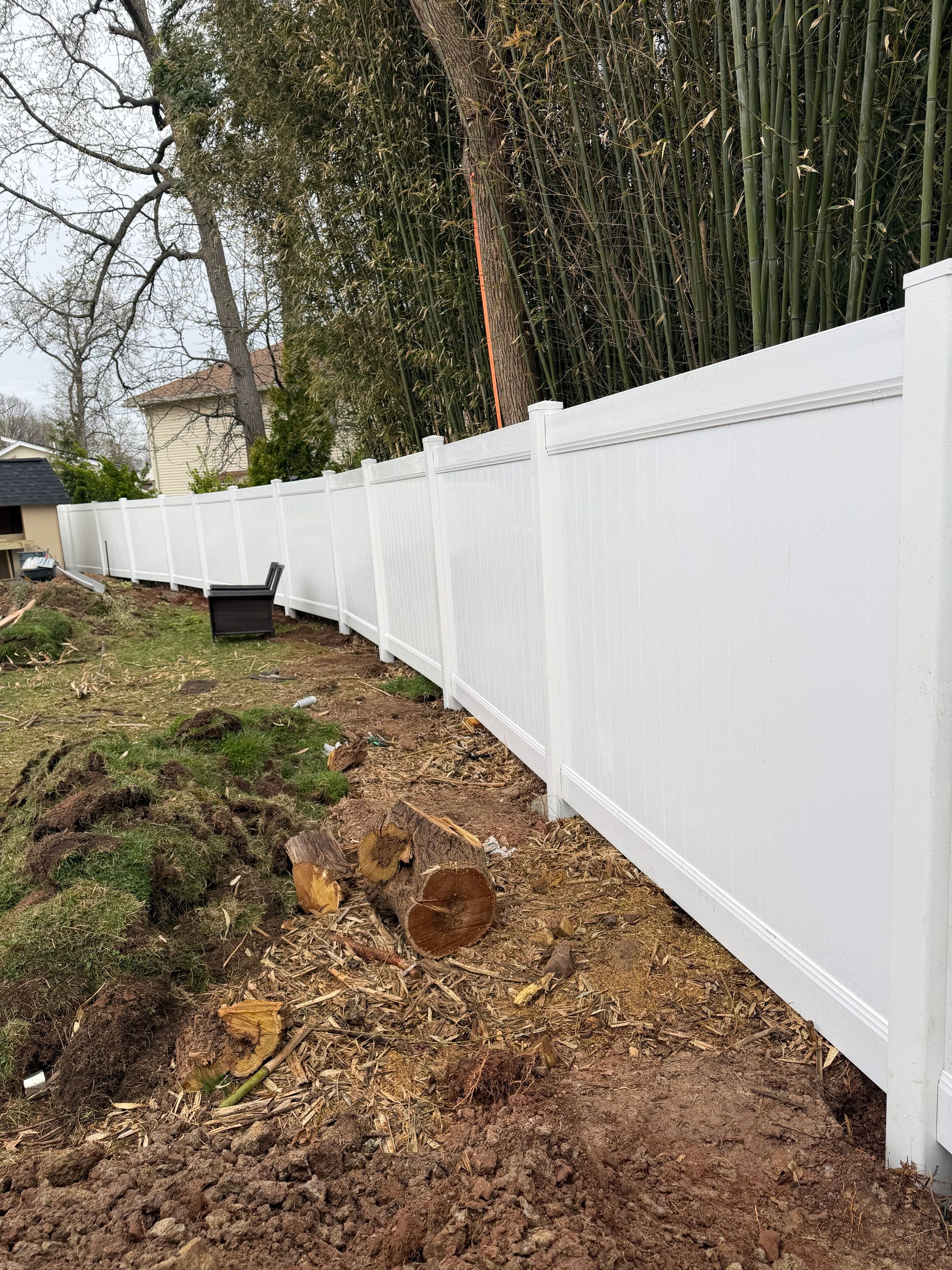 A tall white privacy fence borders a yard with freshly turned dirt and tree stumps, with dense green bamboo in the back.