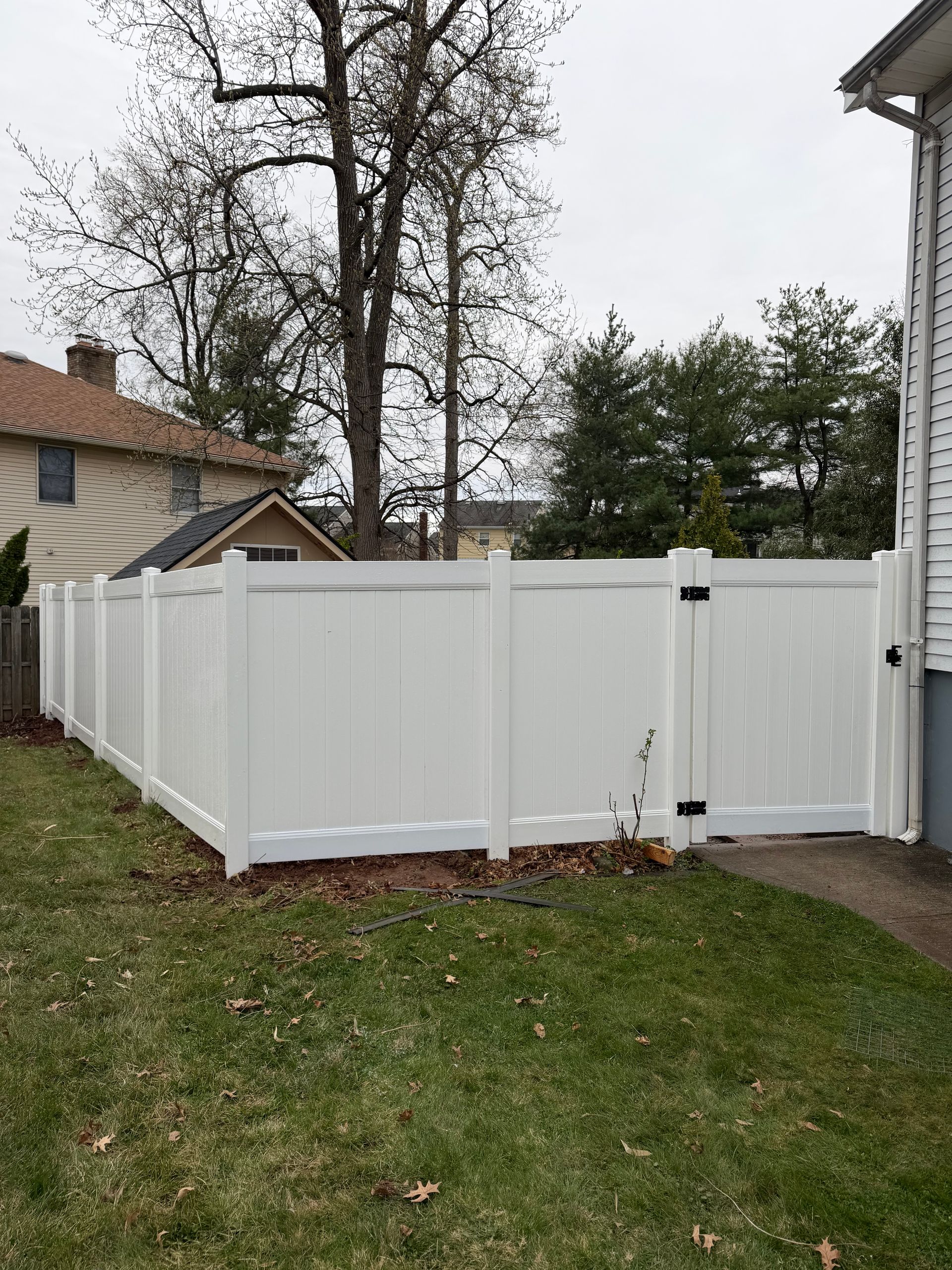 A white vinyl privacy fence with a closed gate extends across a residential backyard lawn on an overcast day.