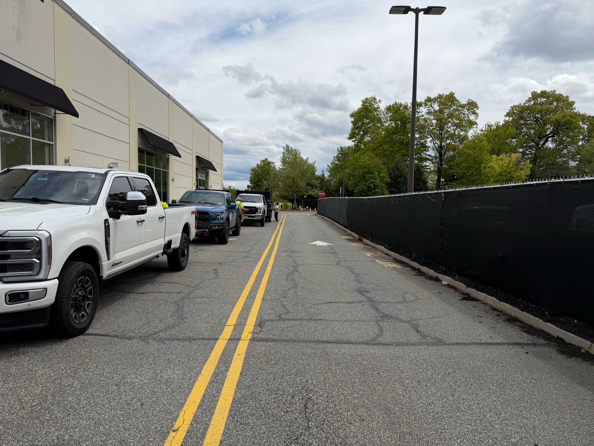 Parking lot lane beside a warehouse, with parked trucks along one side and a black fence on the other.
