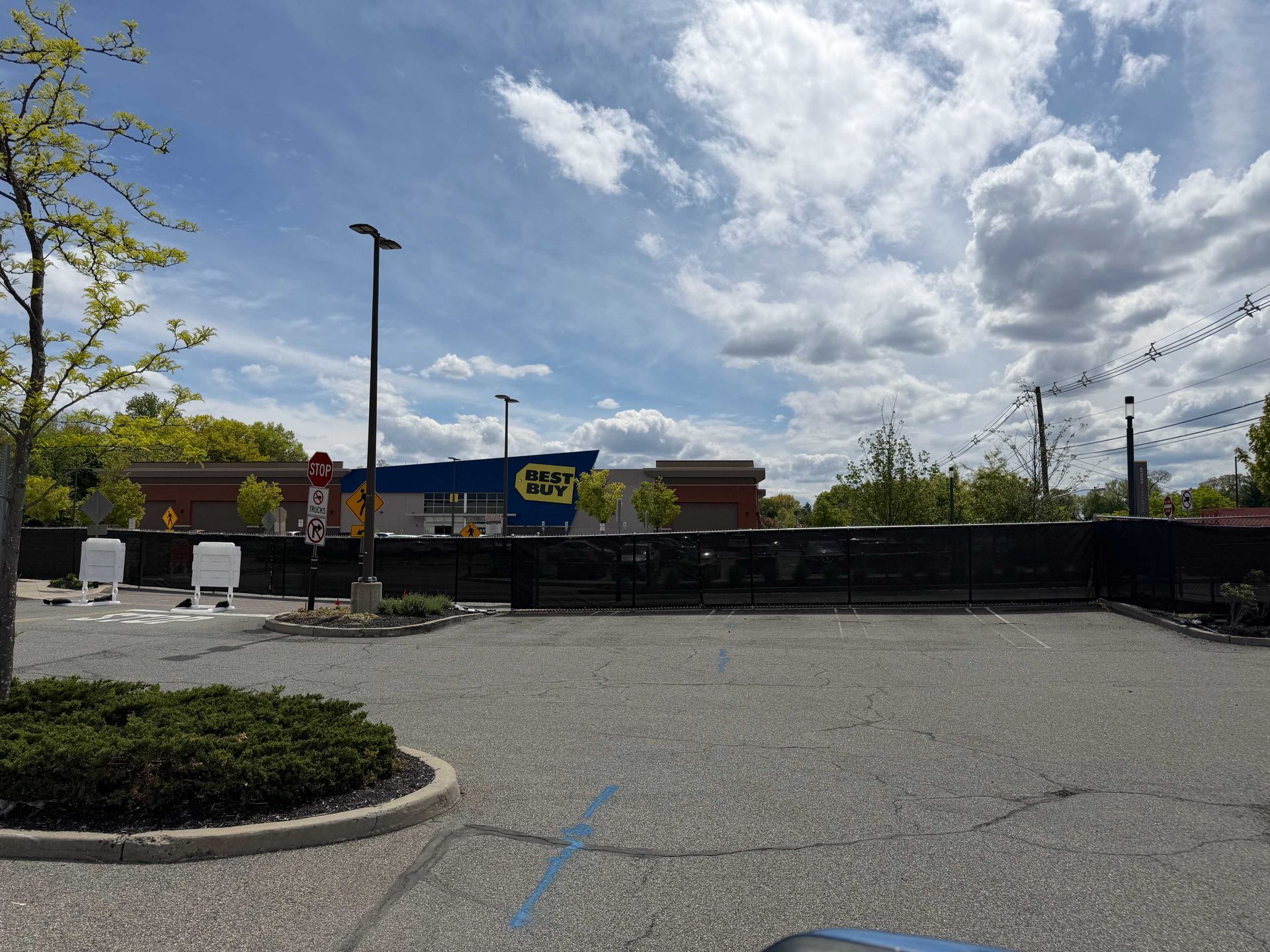 Empty parking lot with a low black fence, storefronts, and a partly cloudy sky.