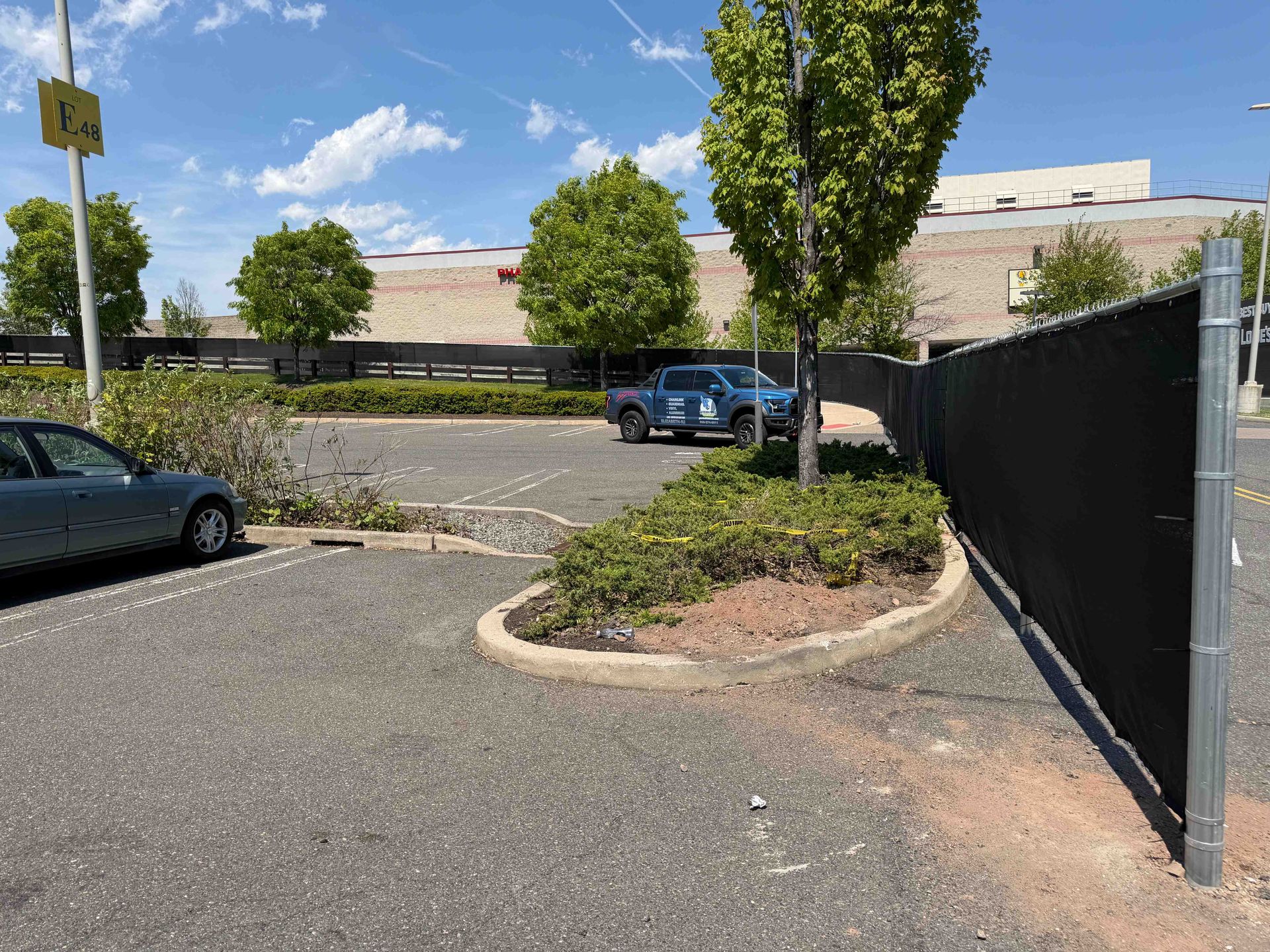 Parking lot with two cars, a tree island, and a black fence beside a beige building under a blue sky