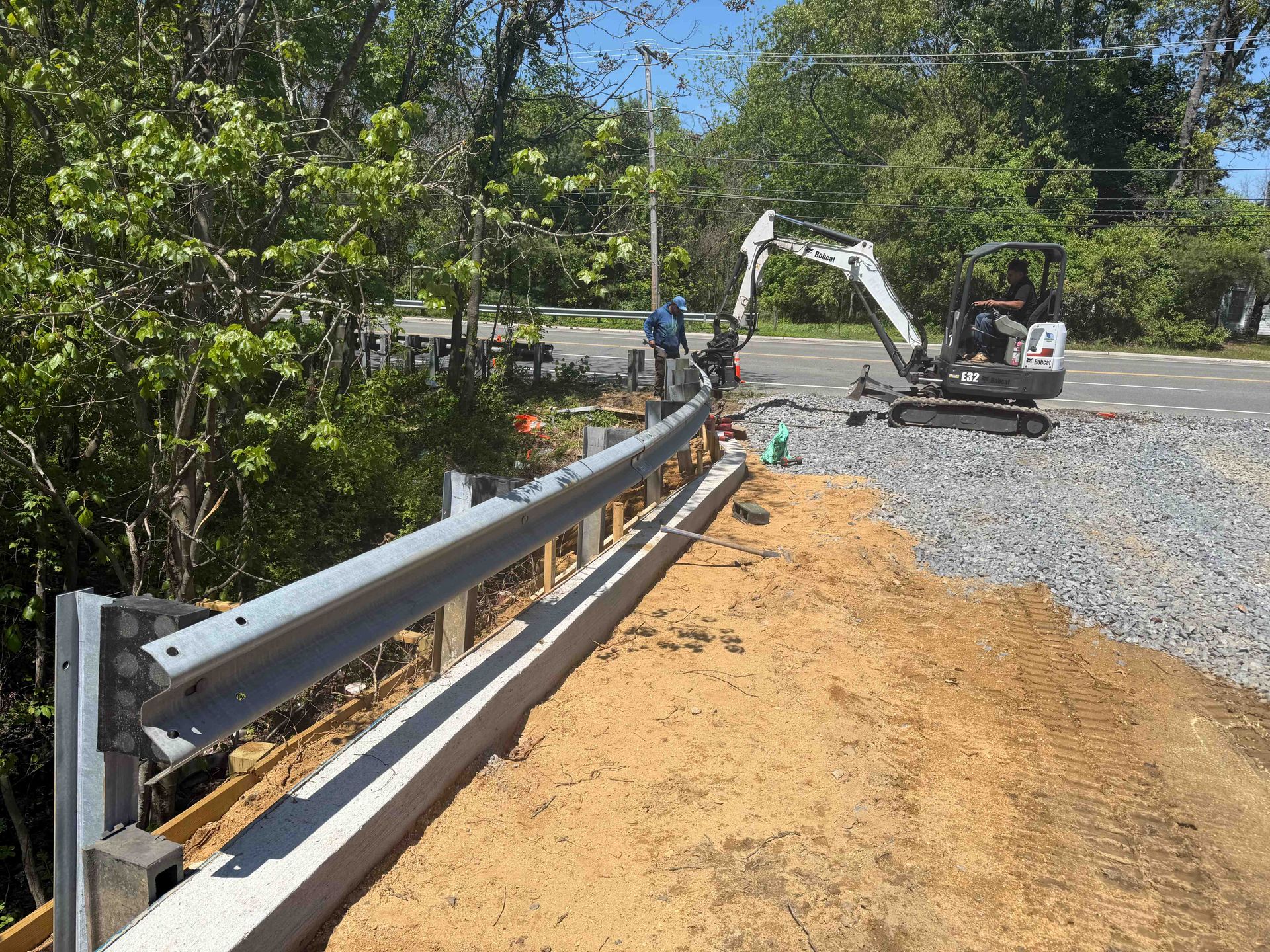 Roadside construction with a guardrail, gravel shoulder, workers, and an excavator under clear skies