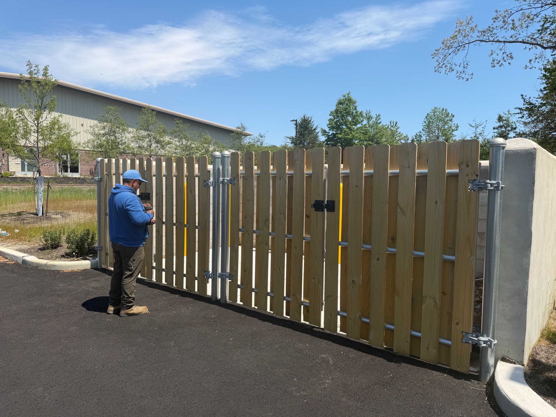 Person in blue jacket opening a tall wooden gate on a paved driveway.