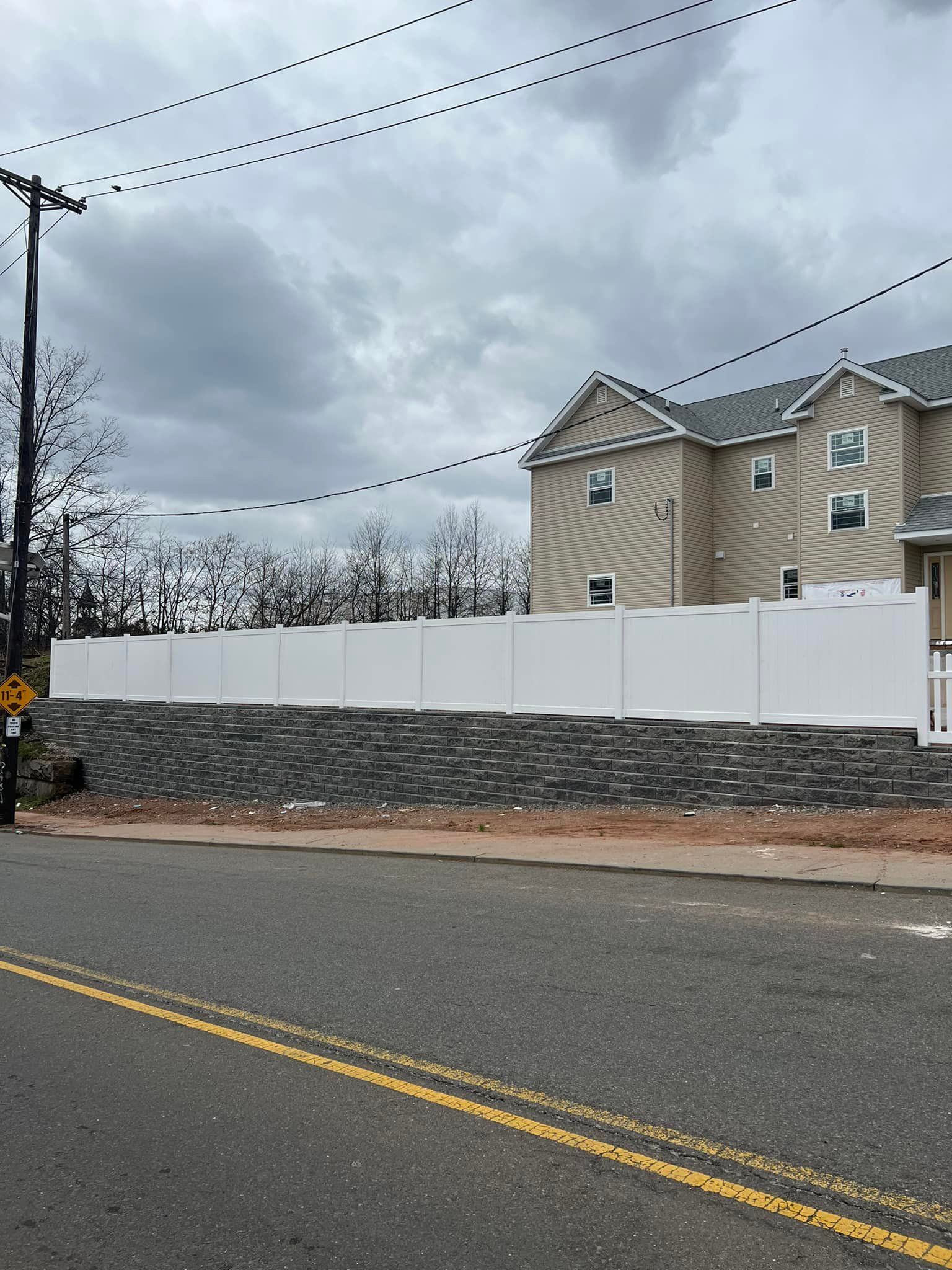 A white fence atop a dark stone retaining wall borders a building along a road under an overcast sky.