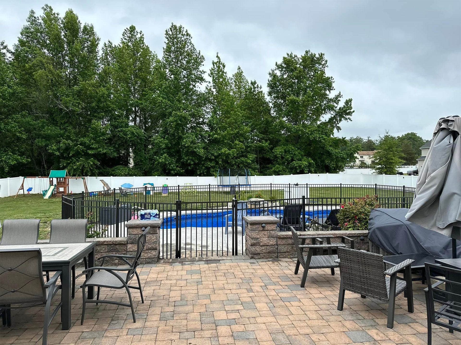 Patio with brick pavers, outdoor furniture, and a fenced-in pool with a green lawn and trees in the background.