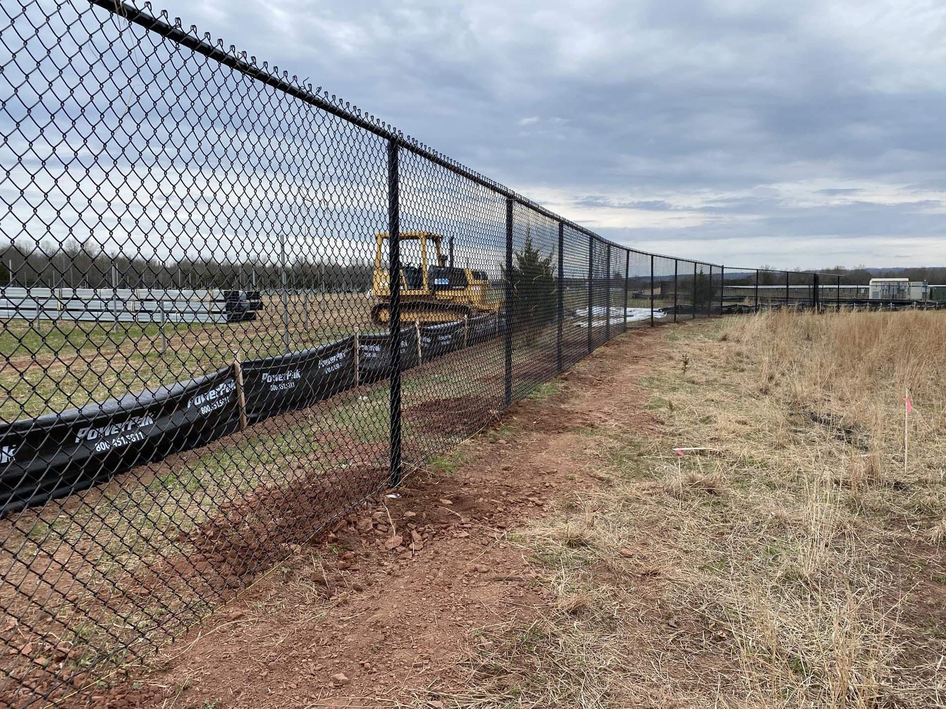 Black chain-link fence bordering a construction site with a bulldozer, dry field, and cloudy sky.