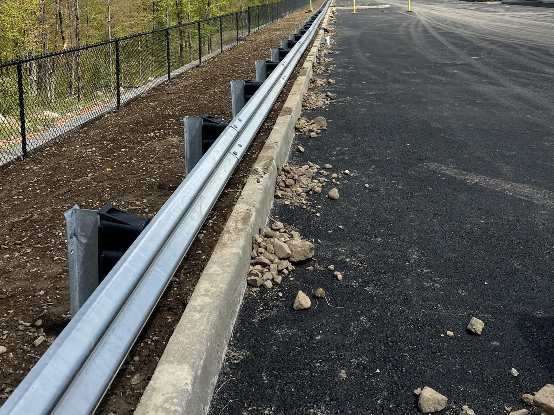 Guardrail next to paved area with black fence and dirt.