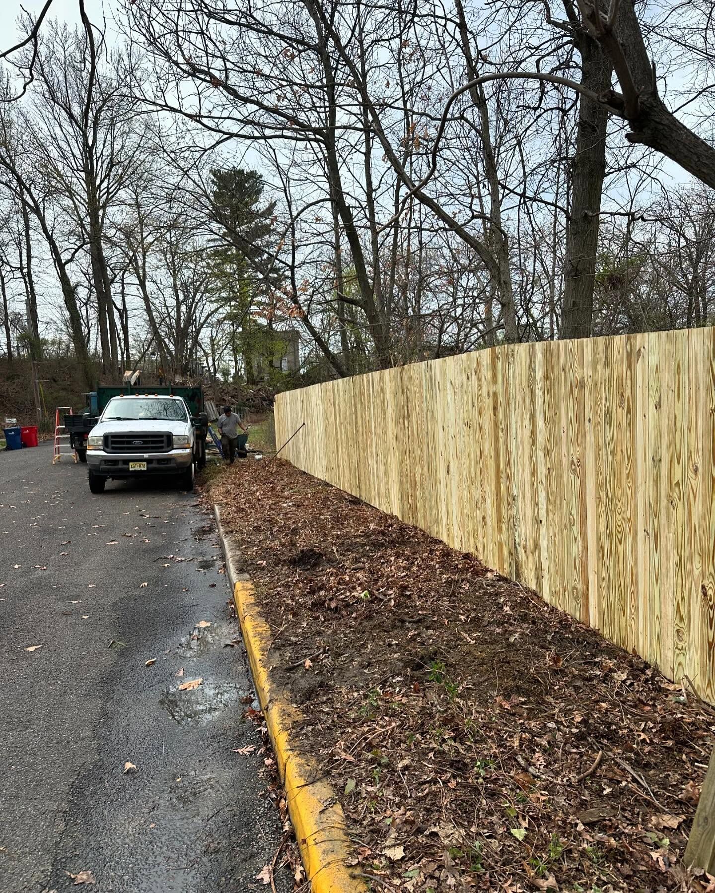 A new wooden fence next to a dirt strip and a paved road with a truck parked. Bare trees in the background.