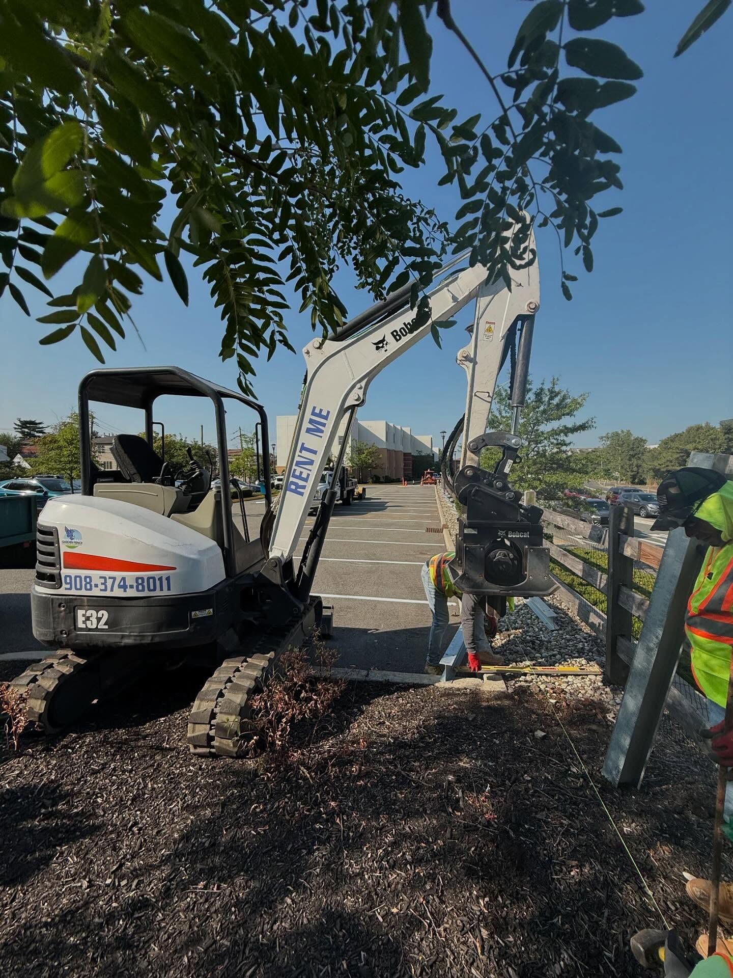 A small Bobcat excavator working next to a wooden fence and pavement, near a tree.