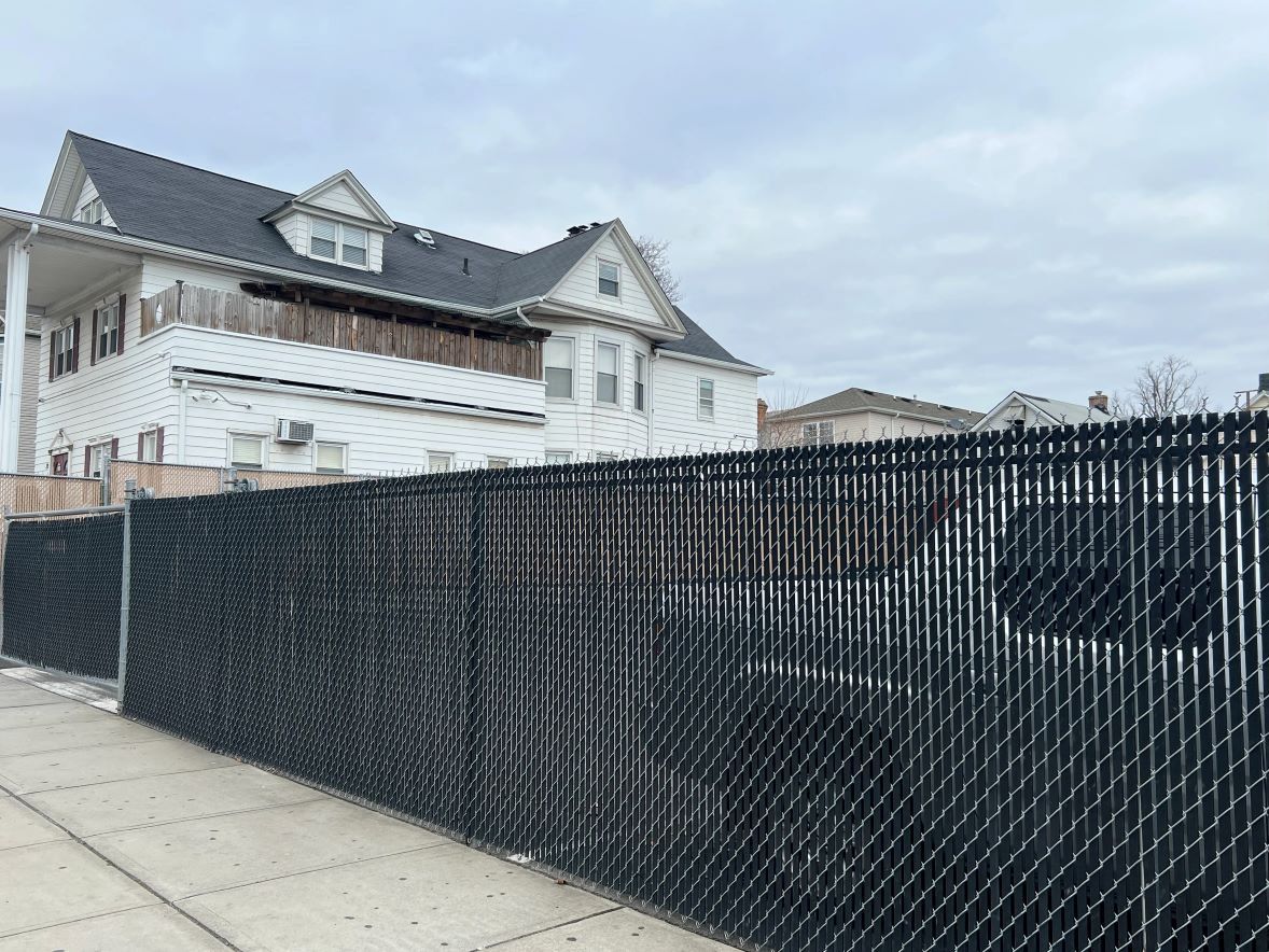 Black chain link fence in front of a white house with a dark roof on a cloudy day.