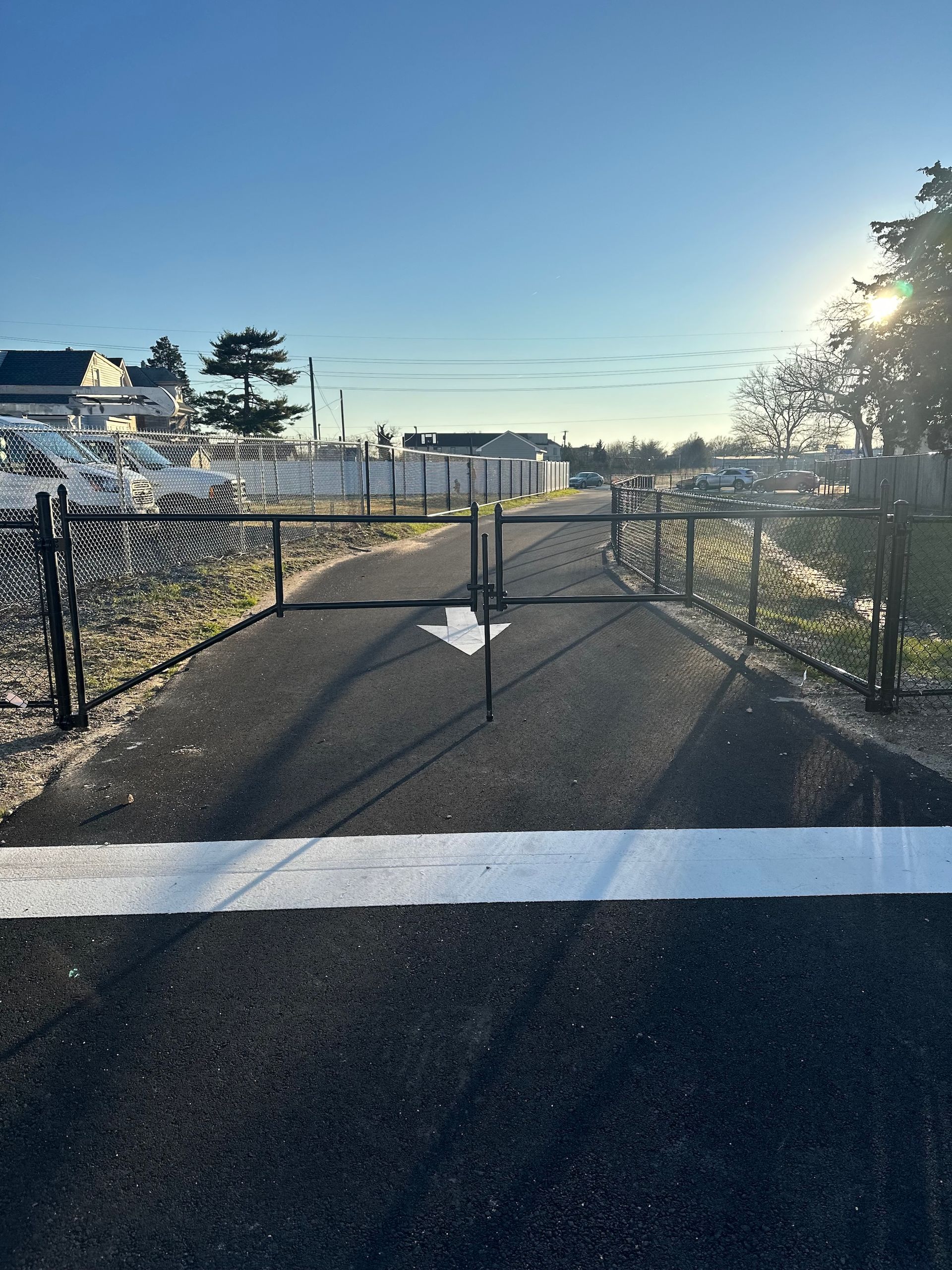 Asphalt path with white arrow leading to a closed black gate. Sunny day with cars in the background.