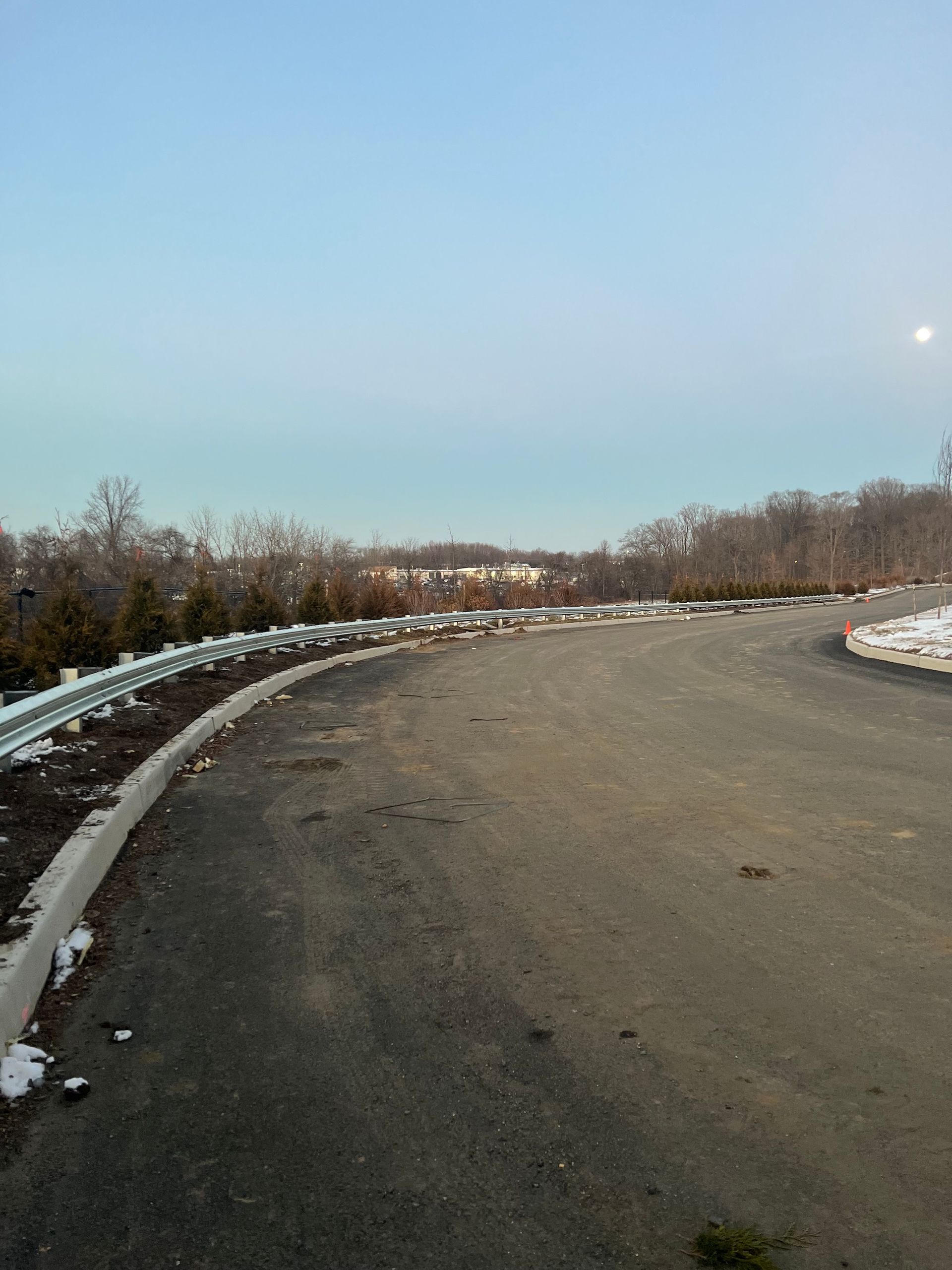 A curving asphalt road with a guardrail and low-lying vegetation under a pale blue sky.