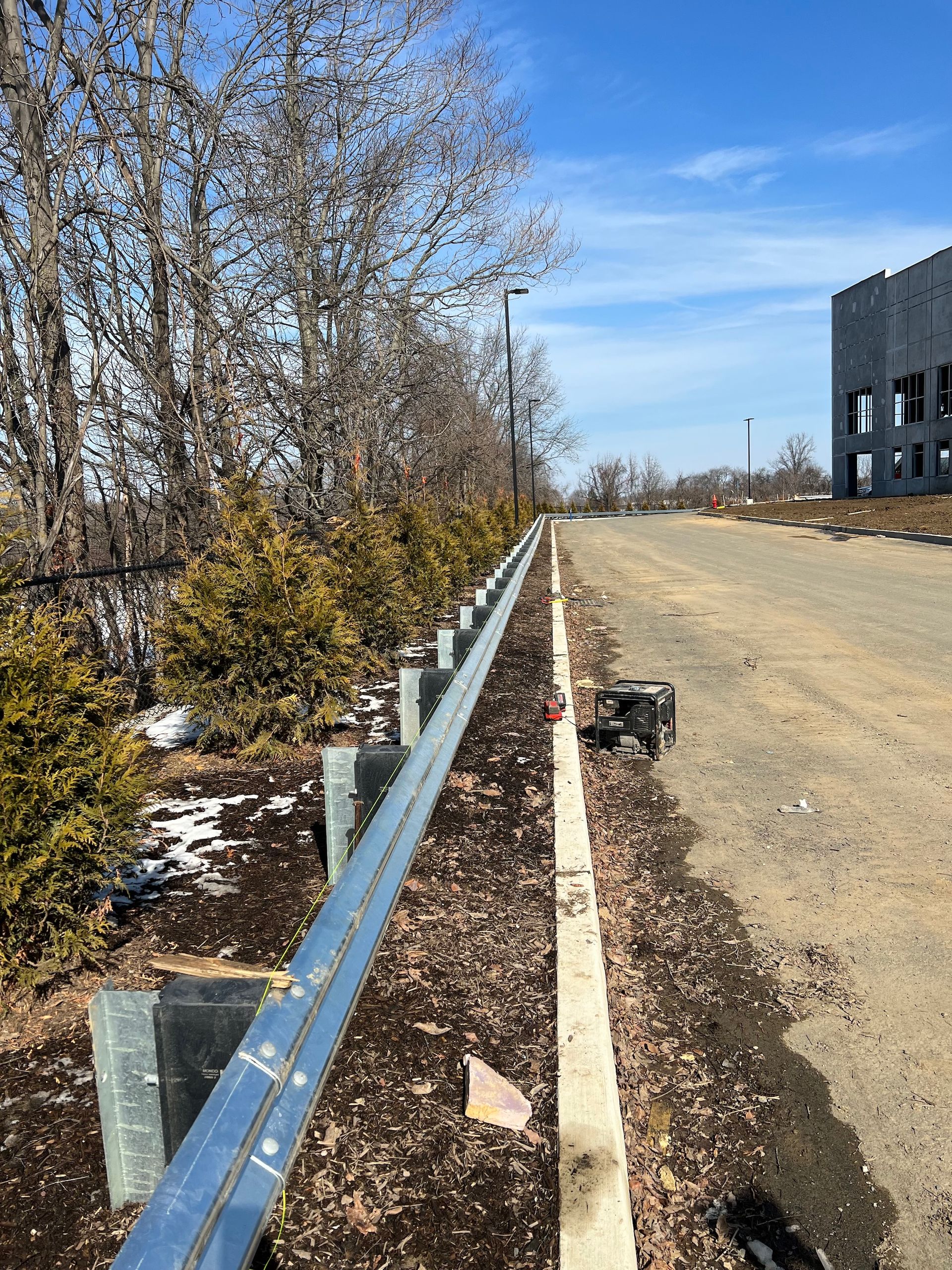 Guardrail alongside a dirt road bordered by evergreen shrubs under a blue sky, with a building under construction visible.