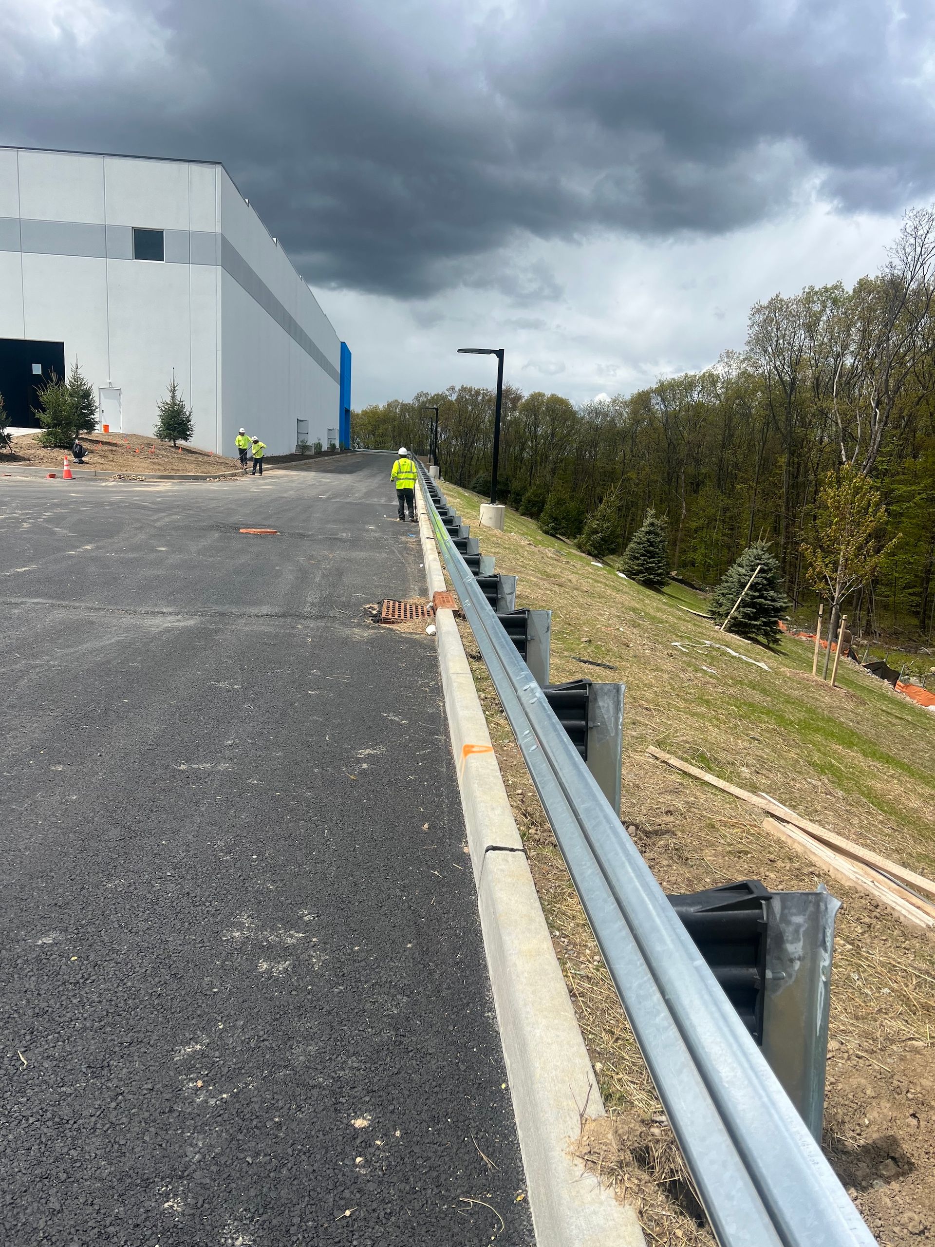 A road with a guardrail next to a grassy slope, with a large building and workers in the background under a cloudy sky.