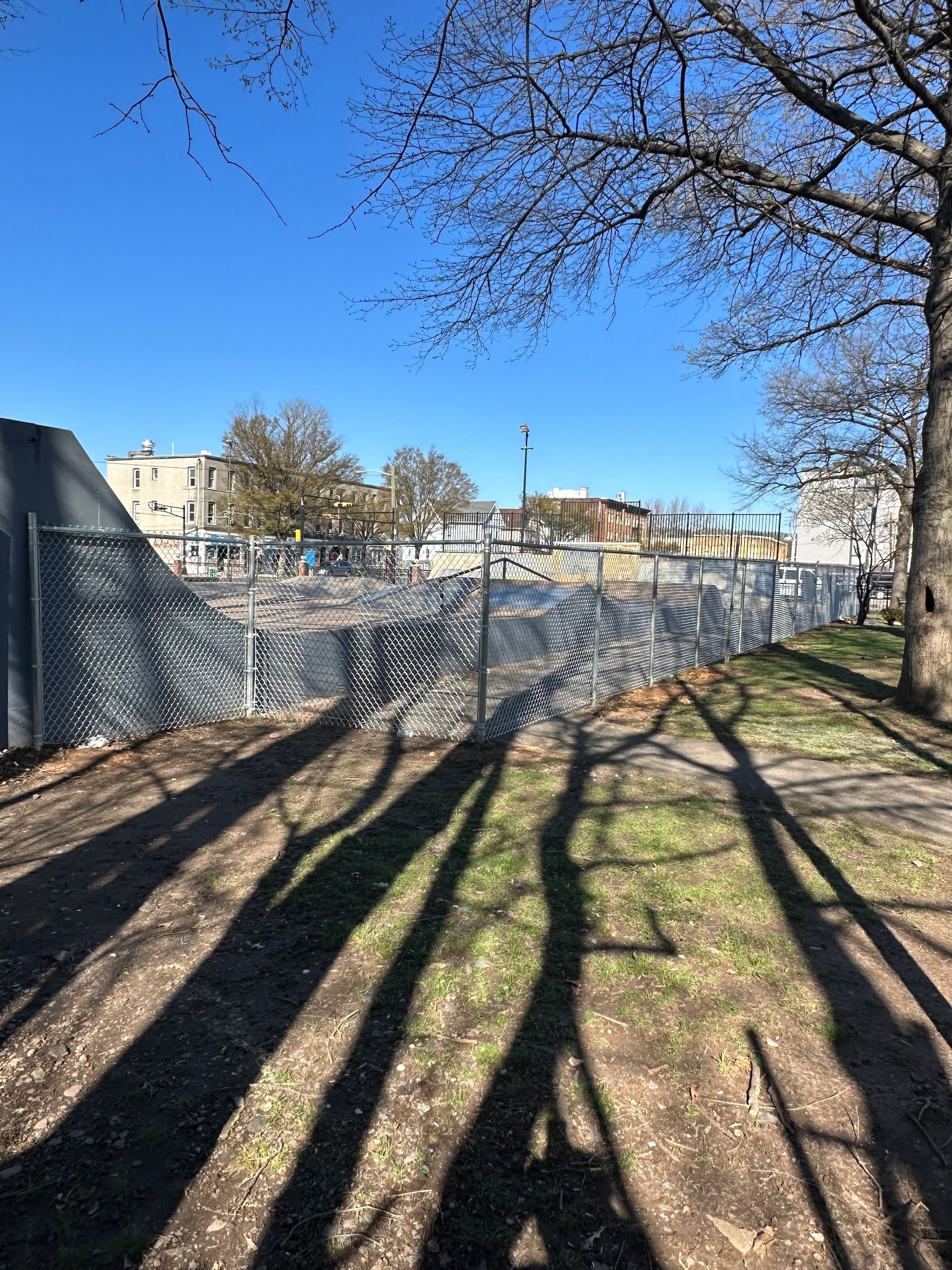 Chain-link fence surrounds a construction site on a sunny day. Long tree shadows cross the grassy ground.