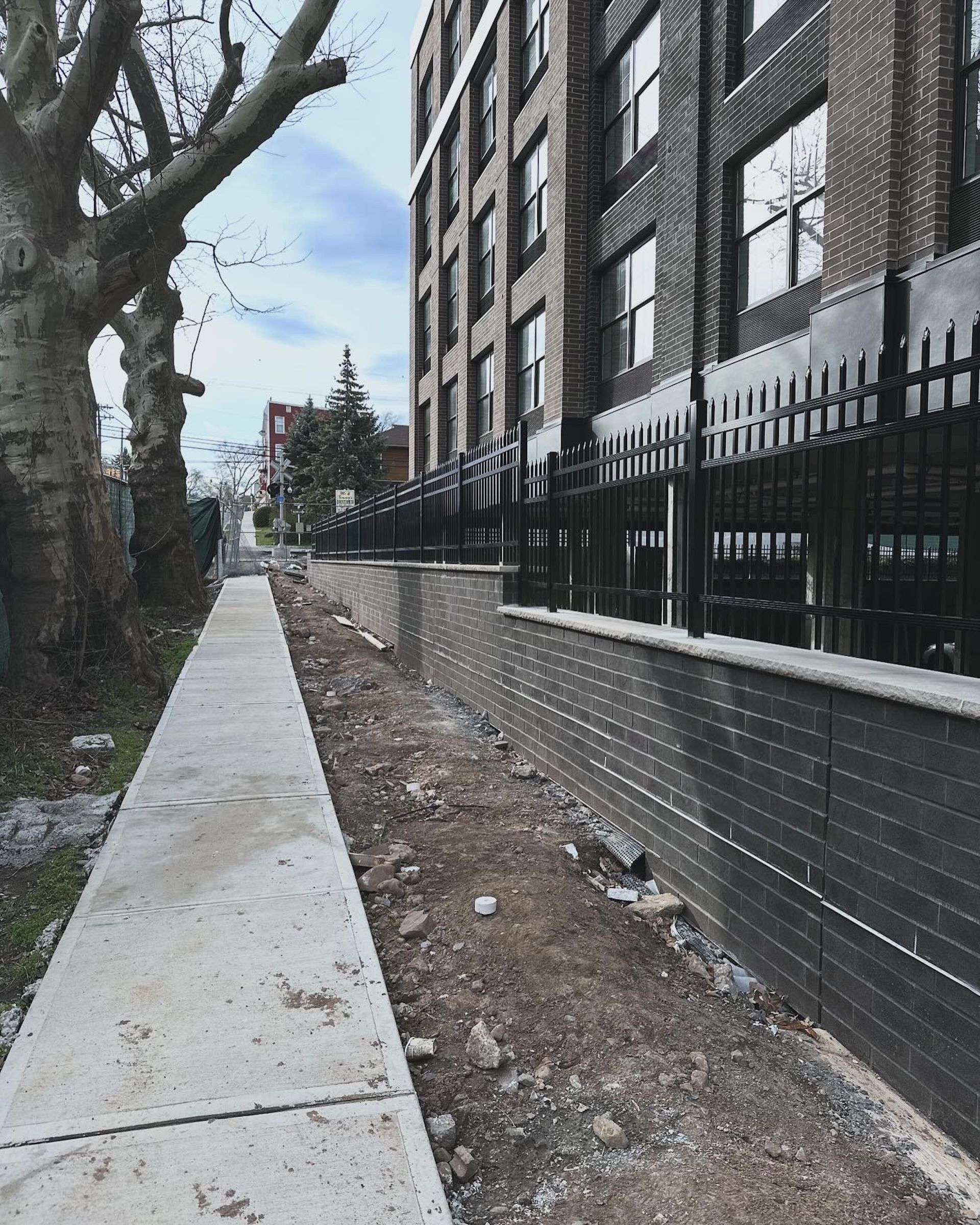 Sidewalk alongside a brick building with a black fence, construction in progress on the right.