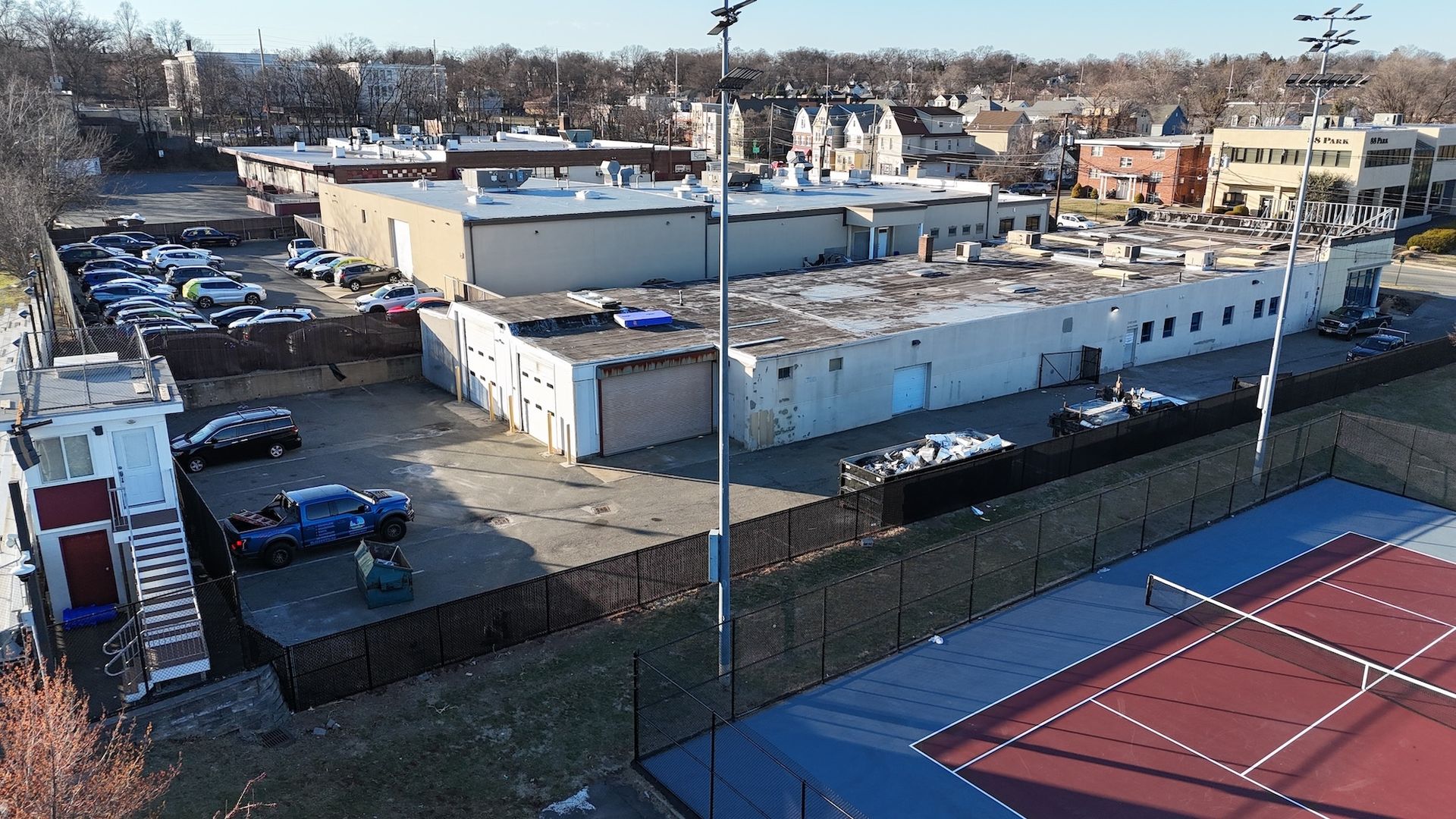 An aerial view shows a commercial area with a tennis court. Gray buildings, parked cars, and trees are visible.