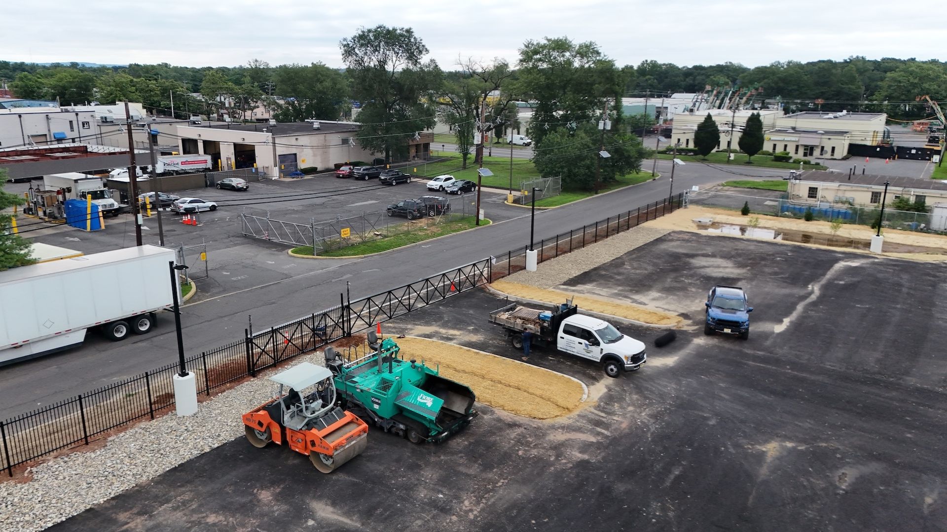 Construction site; paving with machinery; truck, roller, asphalt machine; fenced area; gray sky.