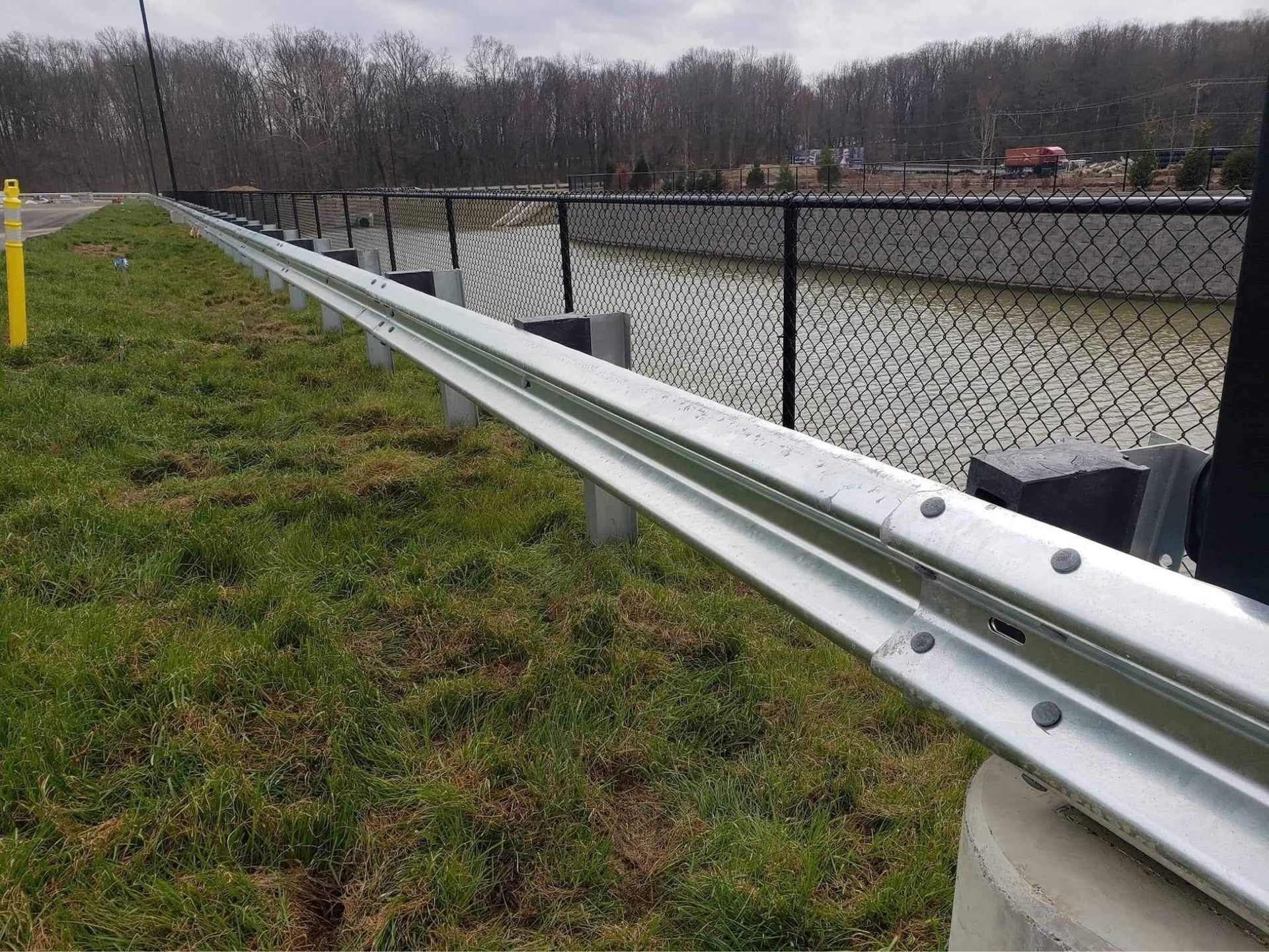 Metal guardrail on grass next to a chain-link fence, trees in background, overcast day.