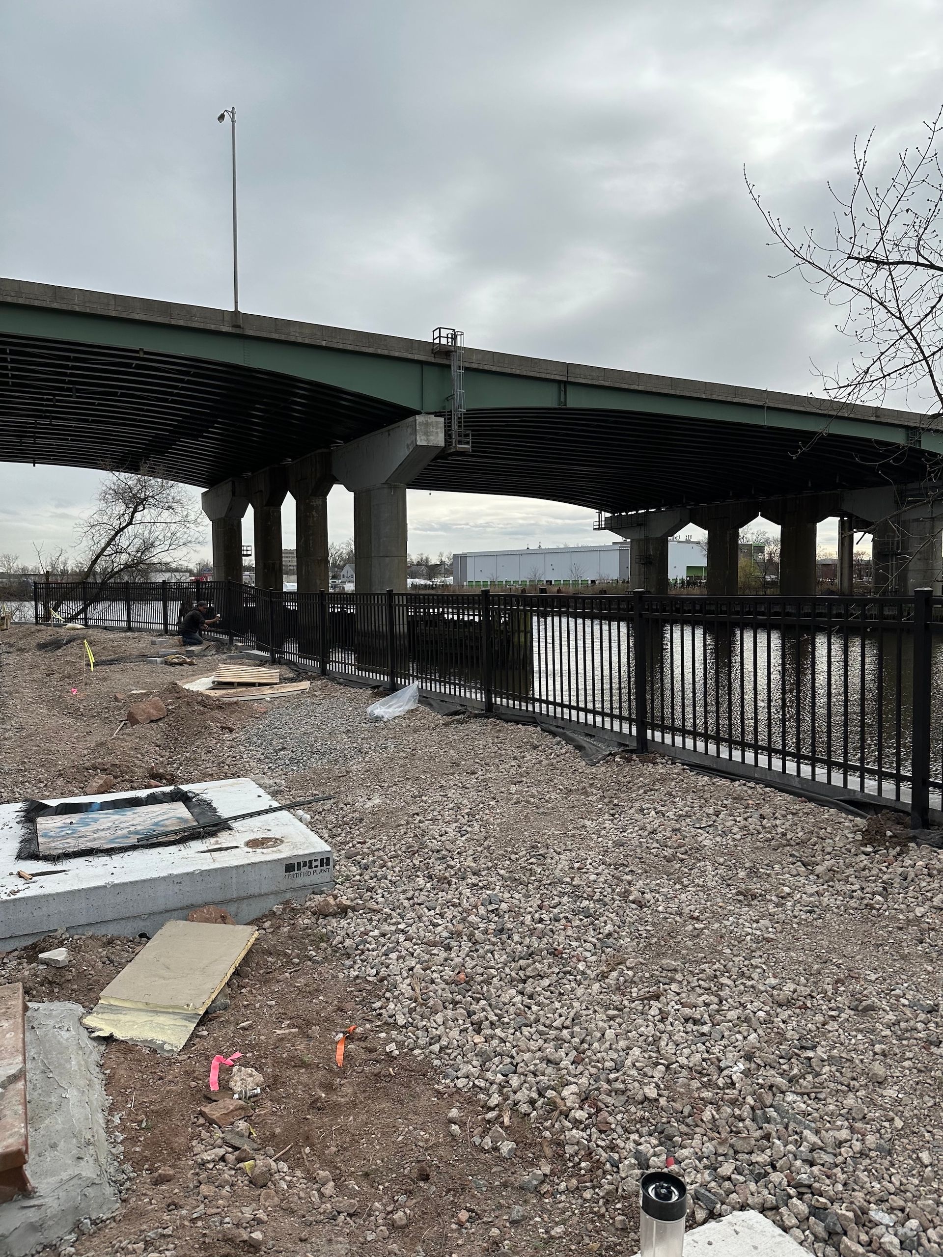 Underneath an elevated highway bridge is a gravel embankment with a black metal fence and water.