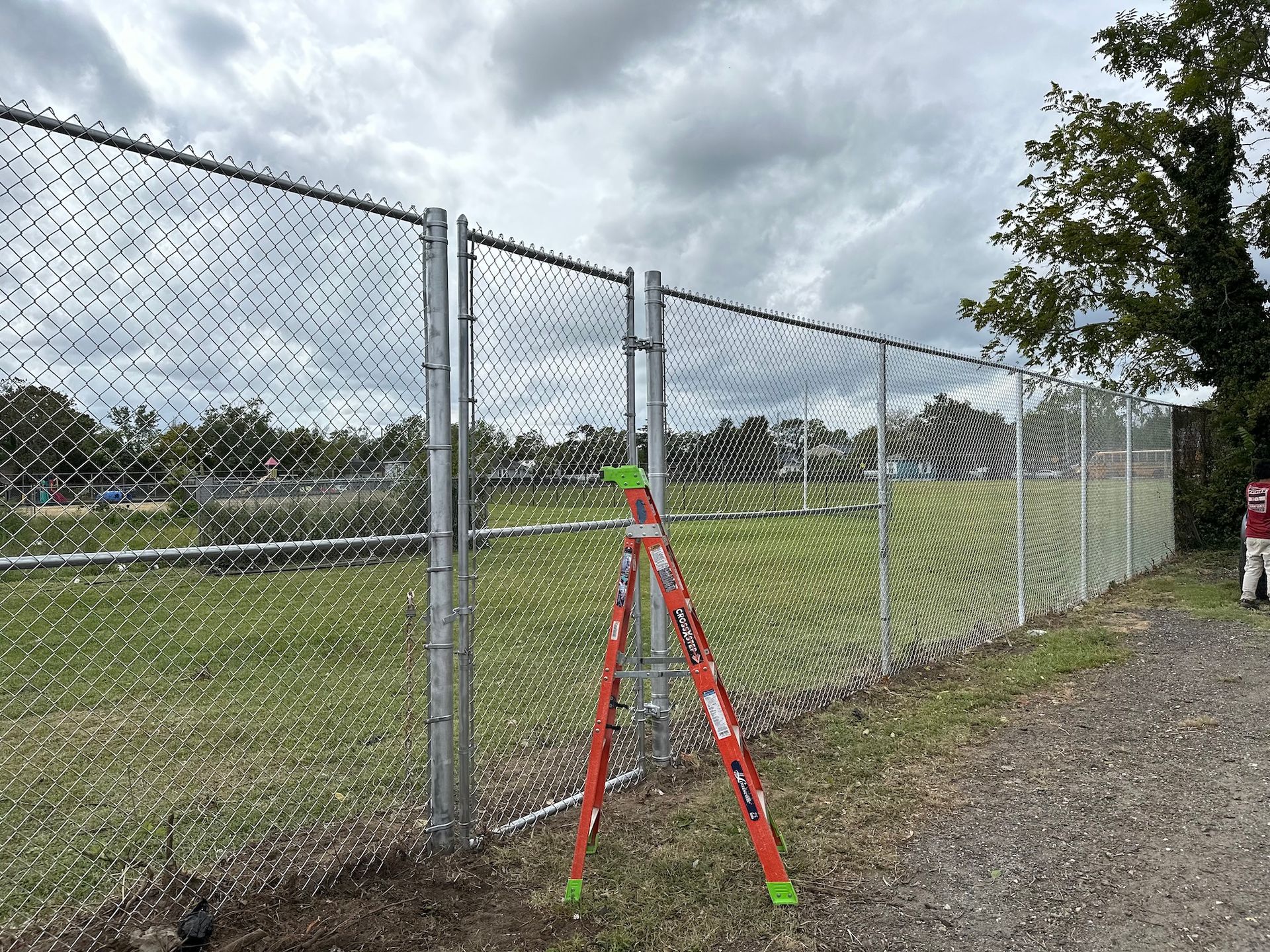 Chain-link fence in front of a grassy field. An orange ladder is leaning against it. Cloudy sky in the background.