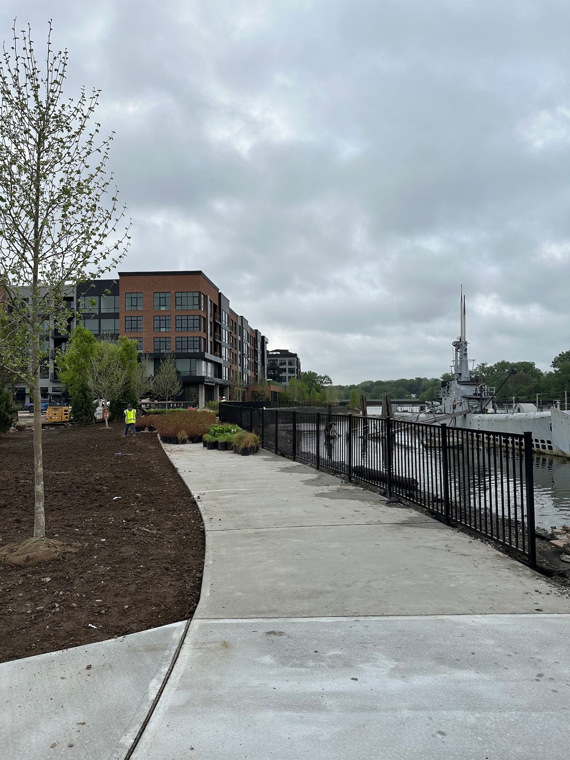 A concrete path curves along a waterfront. Buildings and a submarine are in view. Overcast sky.