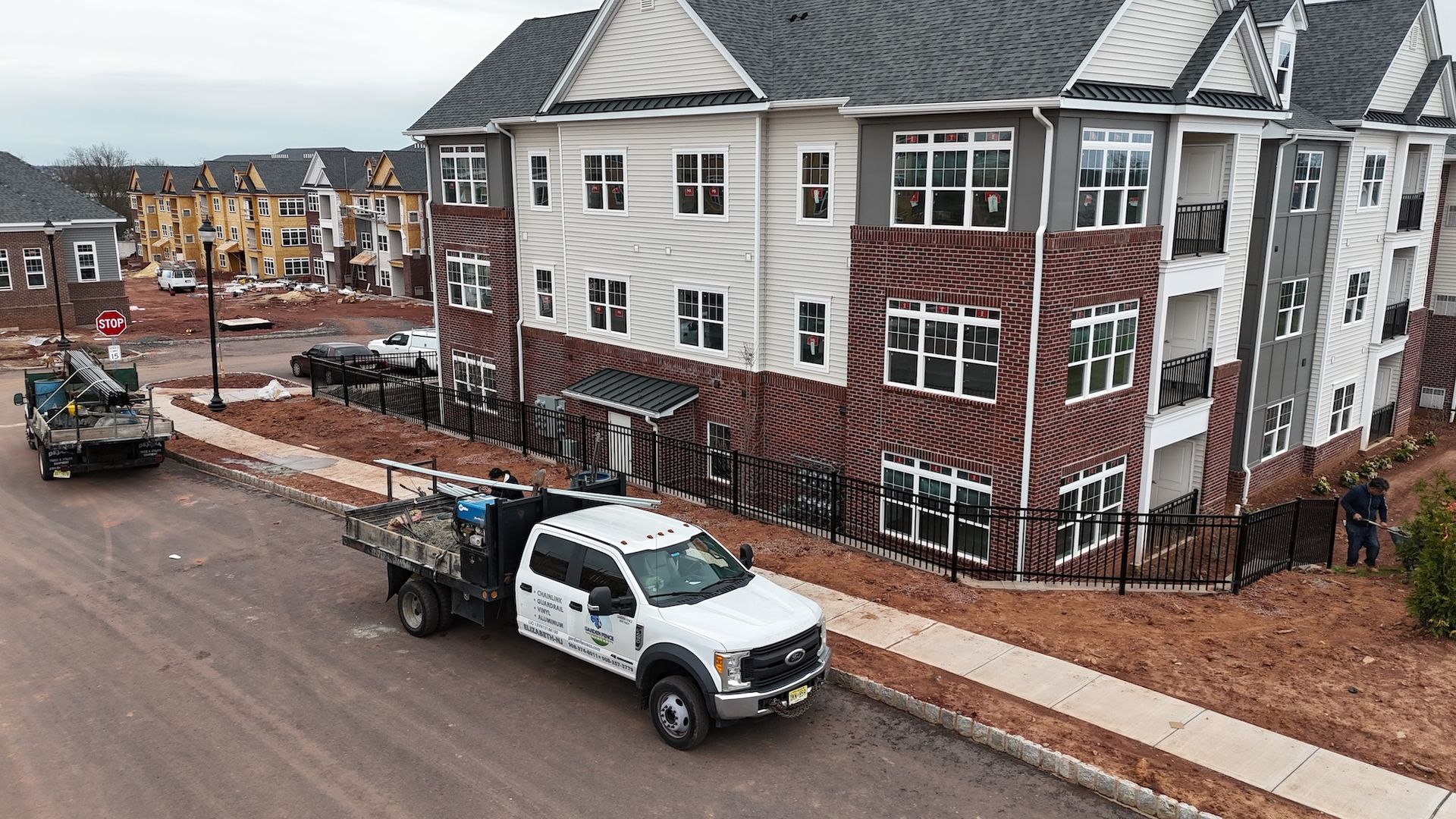 Construction site with multi-story apartment building, work trucks, and a gravel road. Overcast day.
