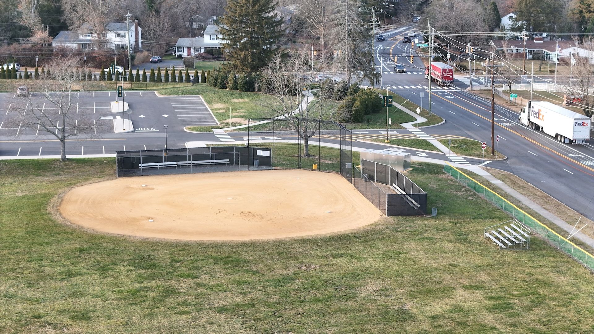 Baseball field in park, next to a road with traffic, a parking lot, and houses.