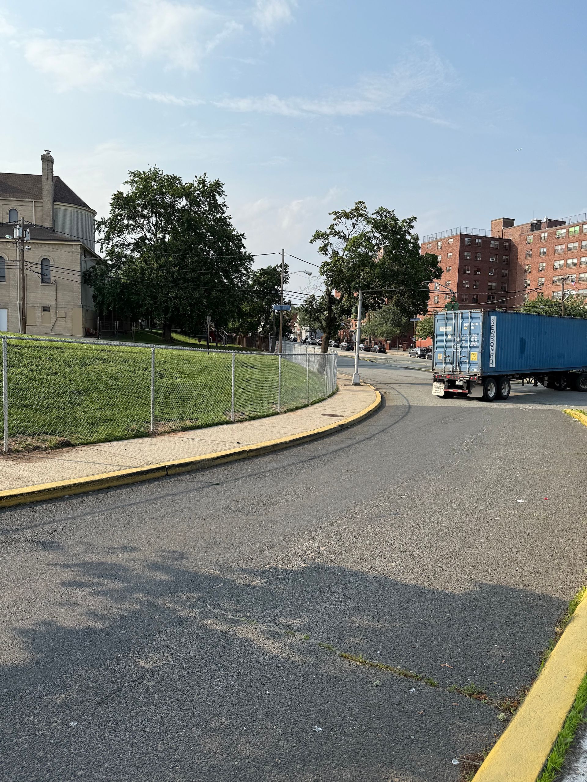 Gravel road with yellow curb leads to a parking area. A blue truck is parked nearby. Green mound, trees, buildings.