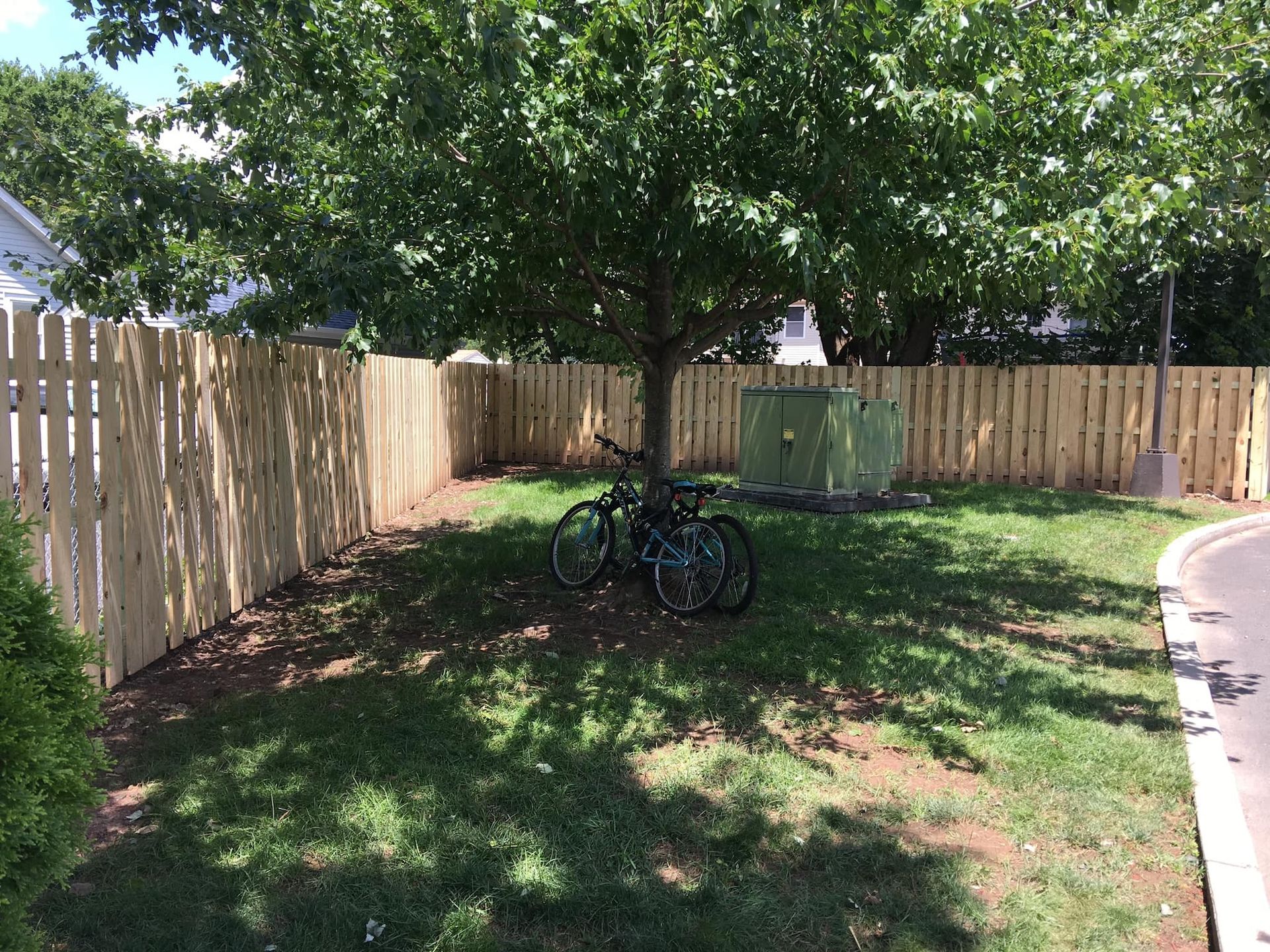 Wooden fence surrounding a grassy area with a tree and two bicycles parked beneath it. A green electrical box is present.