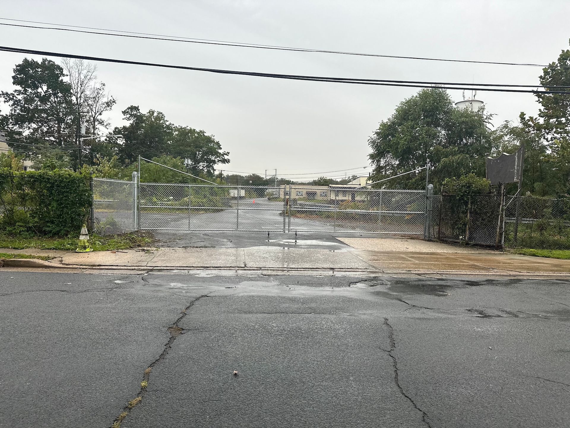 Metal gates block a paved road. A cloudy sky looms. Trees and buildings are visible beyond.