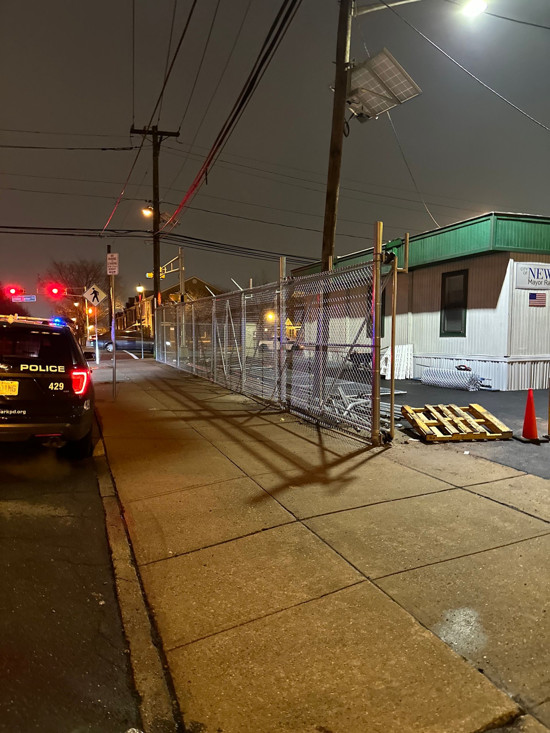 Police car parked on a sidewalk next to a damaged chain-link fence at night.