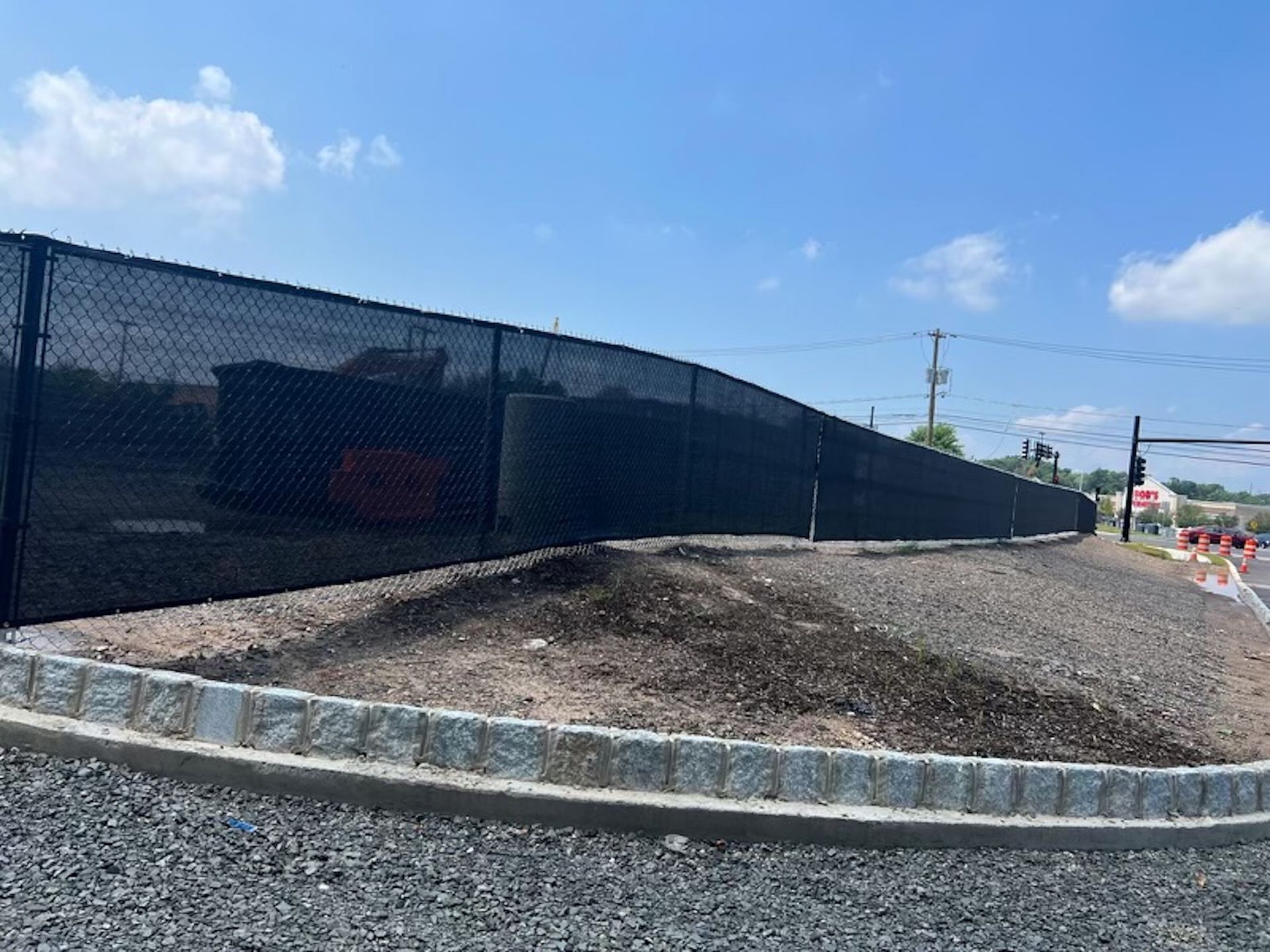 Black chain-link fence with privacy screening surrounds a construction site; blue sky overhead.
