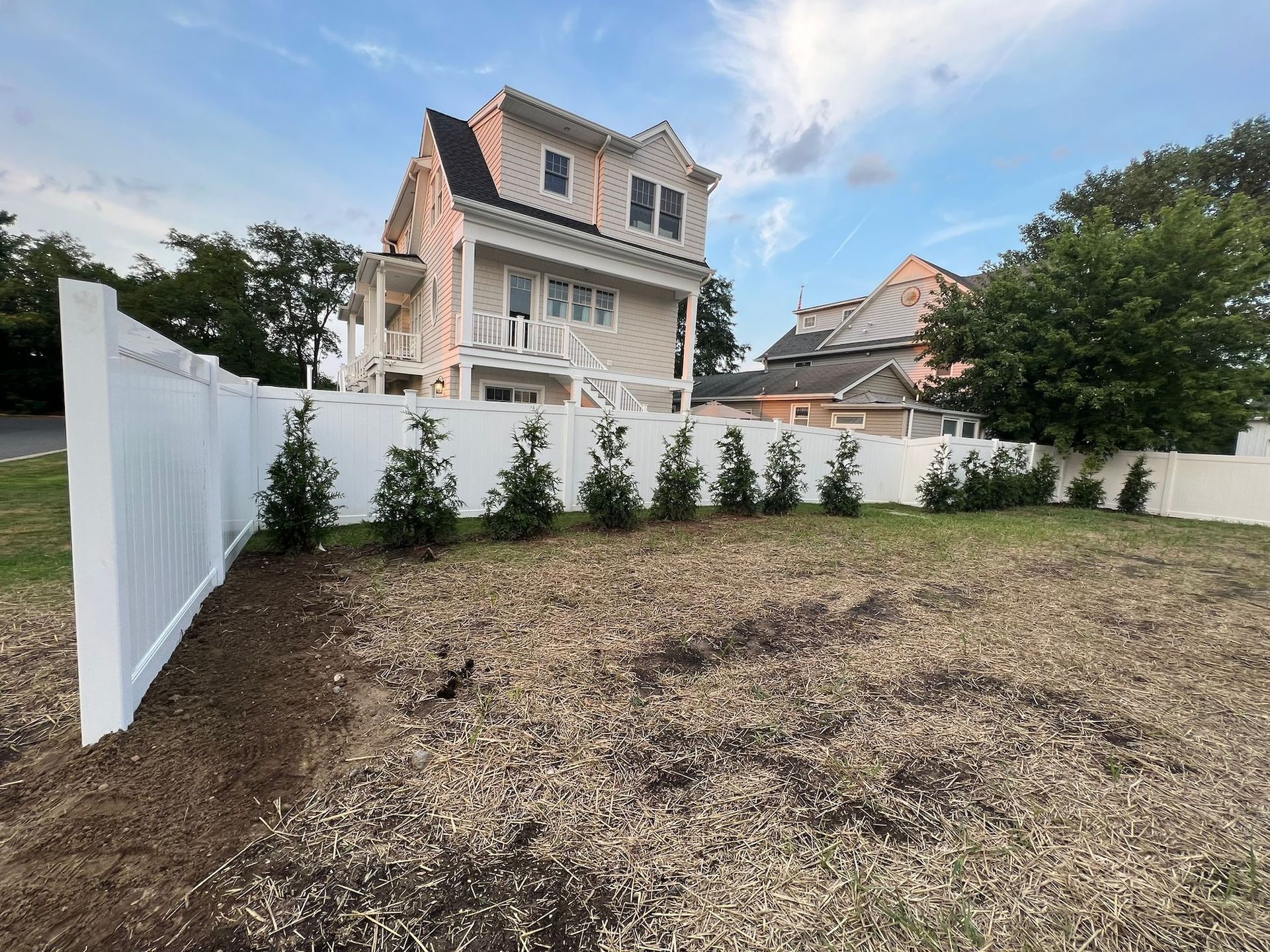 White fence and newly planted small trees in front of a light-colored house with a deck under a cloudy sky.