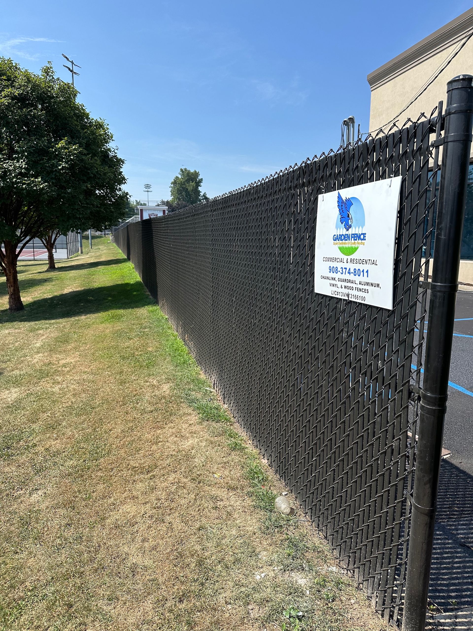 Black chain-link fence with a sign, next to a grassy area and building, blue sky.